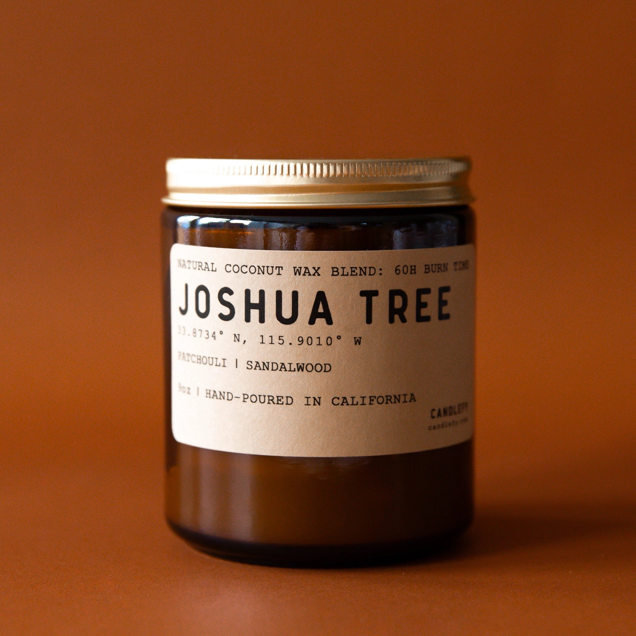  An amber colored glass candle jar with a brown label on the front that reads, "Joshua Tree Patchouli | Sandalwood" in black letter along with a single wick candle inside the jar and a gold screw on lid.