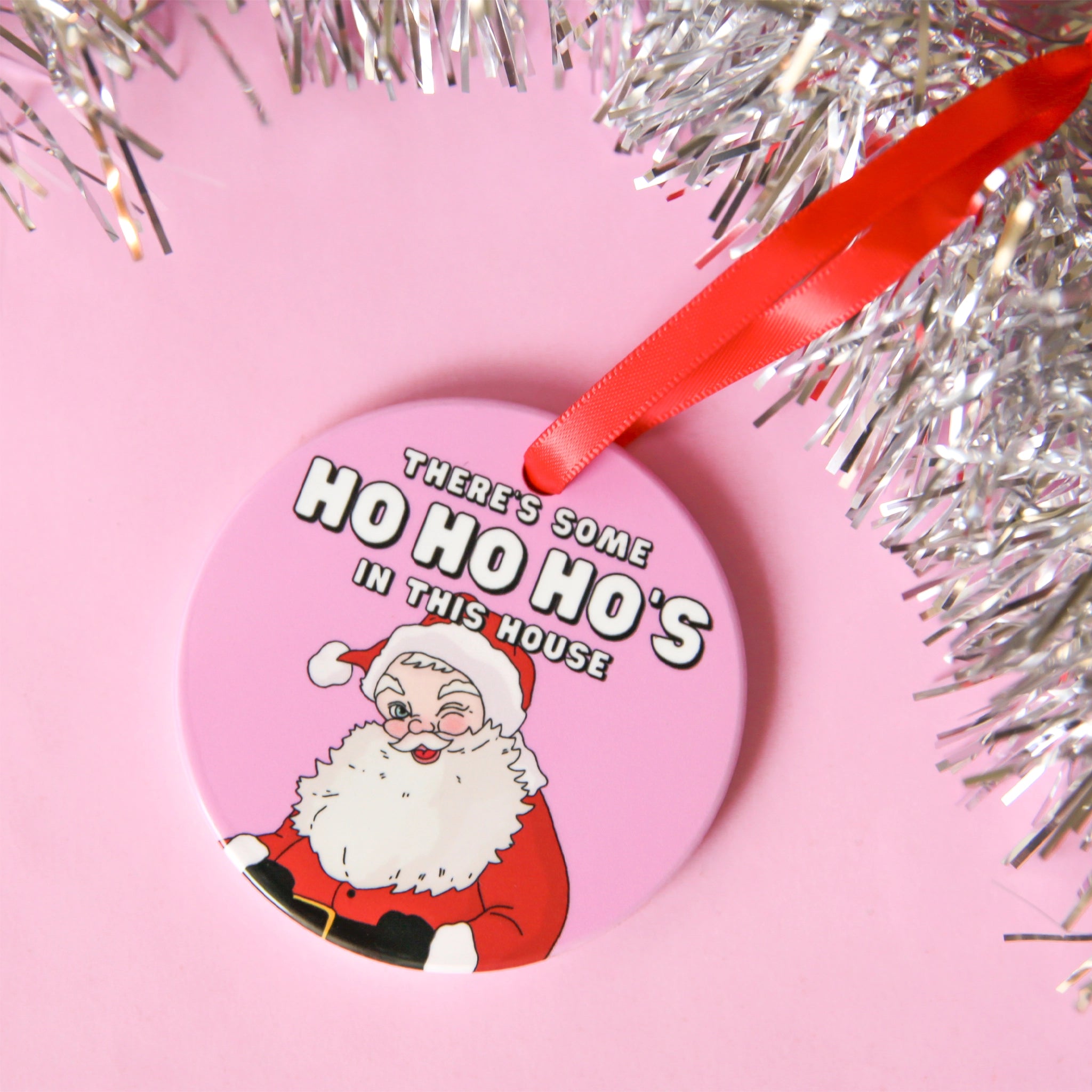 On a pink background is a round pink ornament with a graphic of Santa winking and white text above that reads, &quot;There&#39;s Some Ho Ho Ho&#39;s In This House&quot; and a red ribbon loop for hanging.