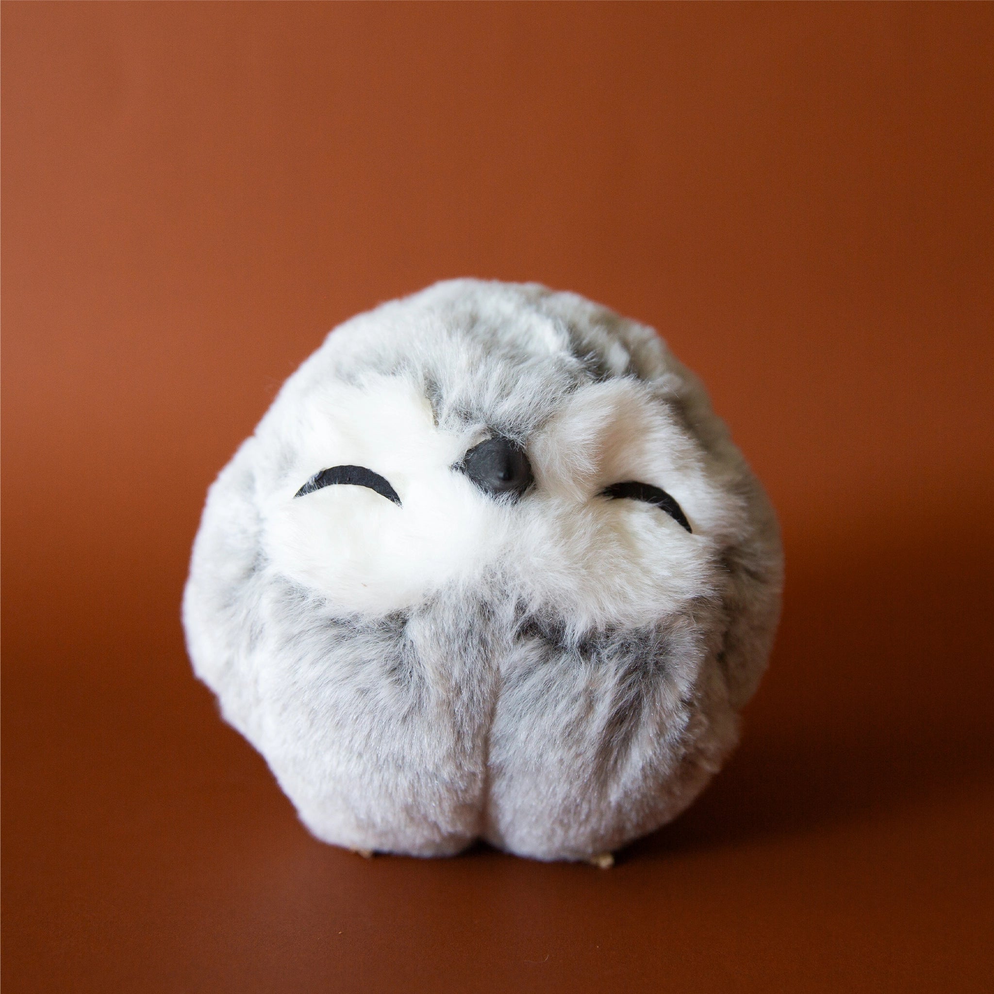 On a brown background is a grey an white faux fur ornament with closed eyes. 