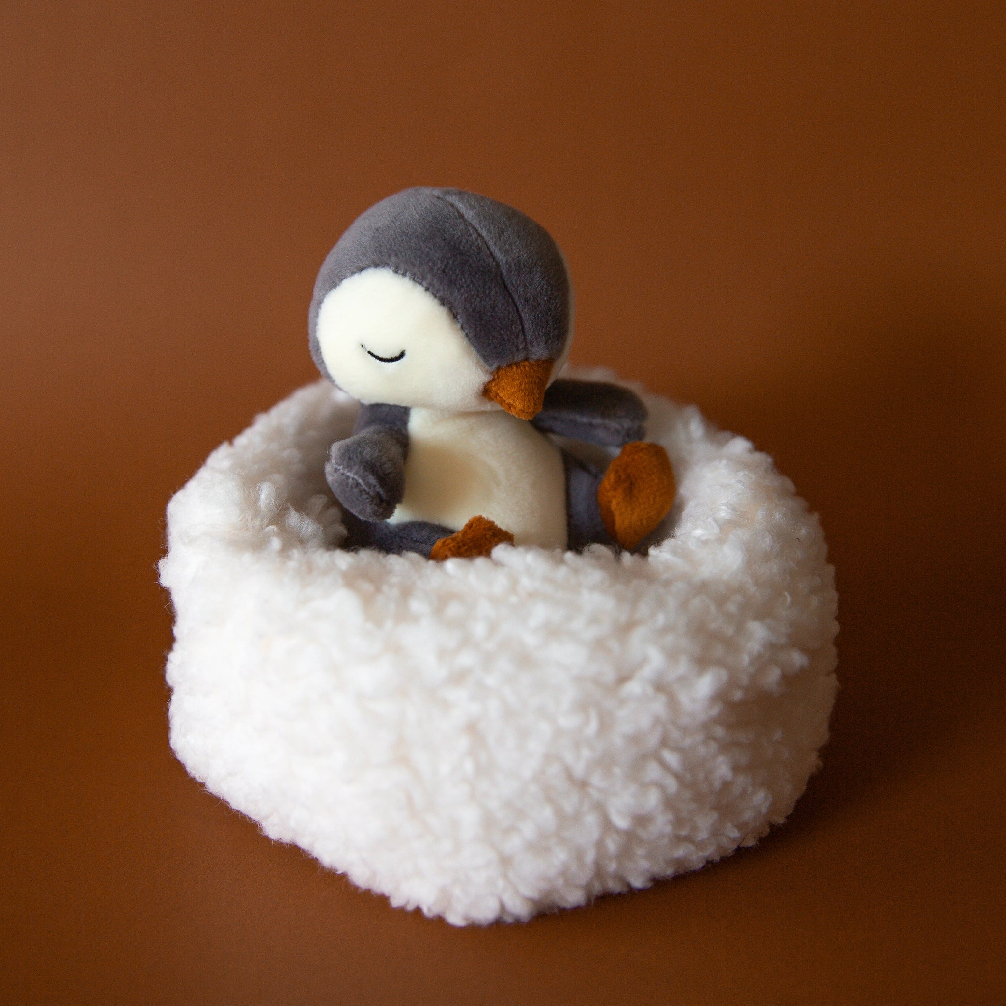 On a brown background is a penguin stuffed animal with a white boucle bed.