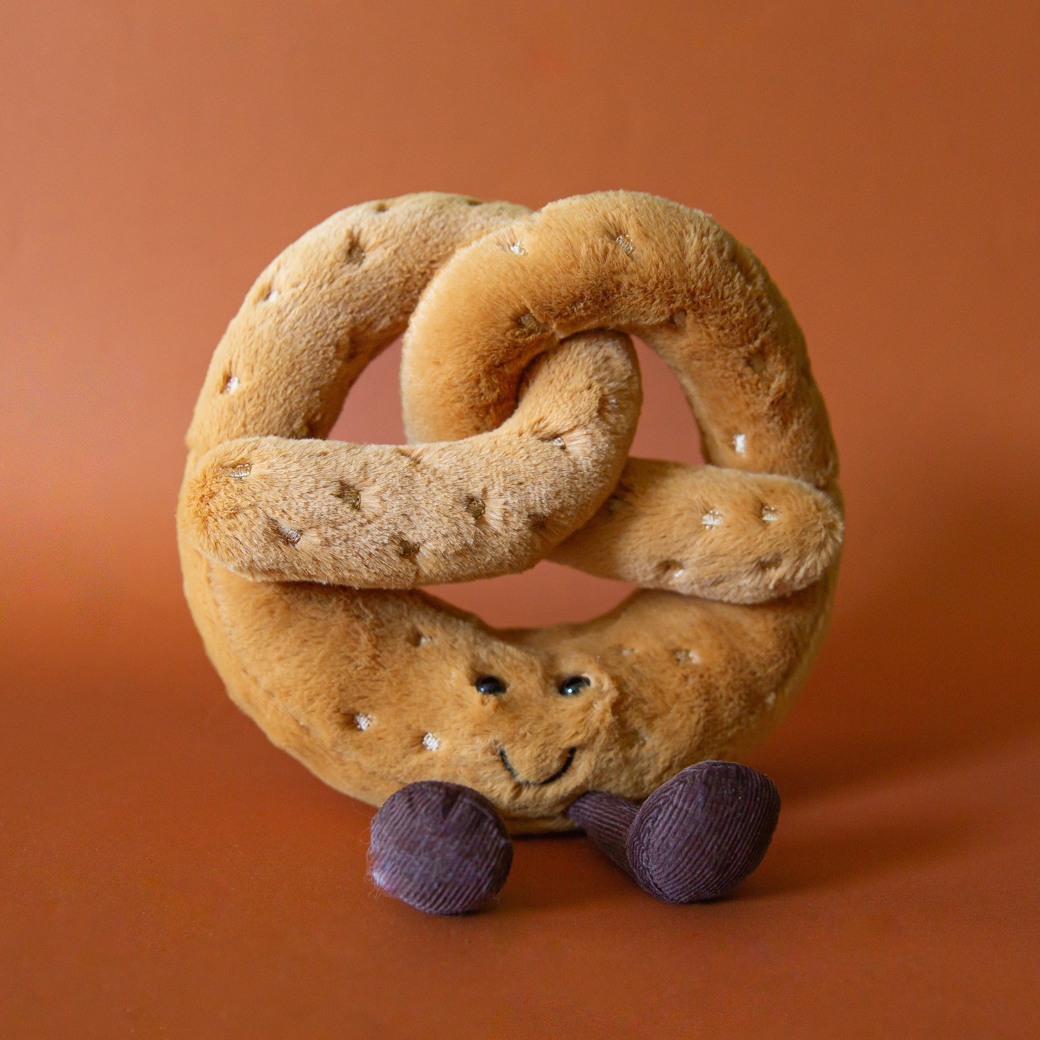 On a burnt orange background is a tan pretzel stuffed toy with a smiling expression. 
