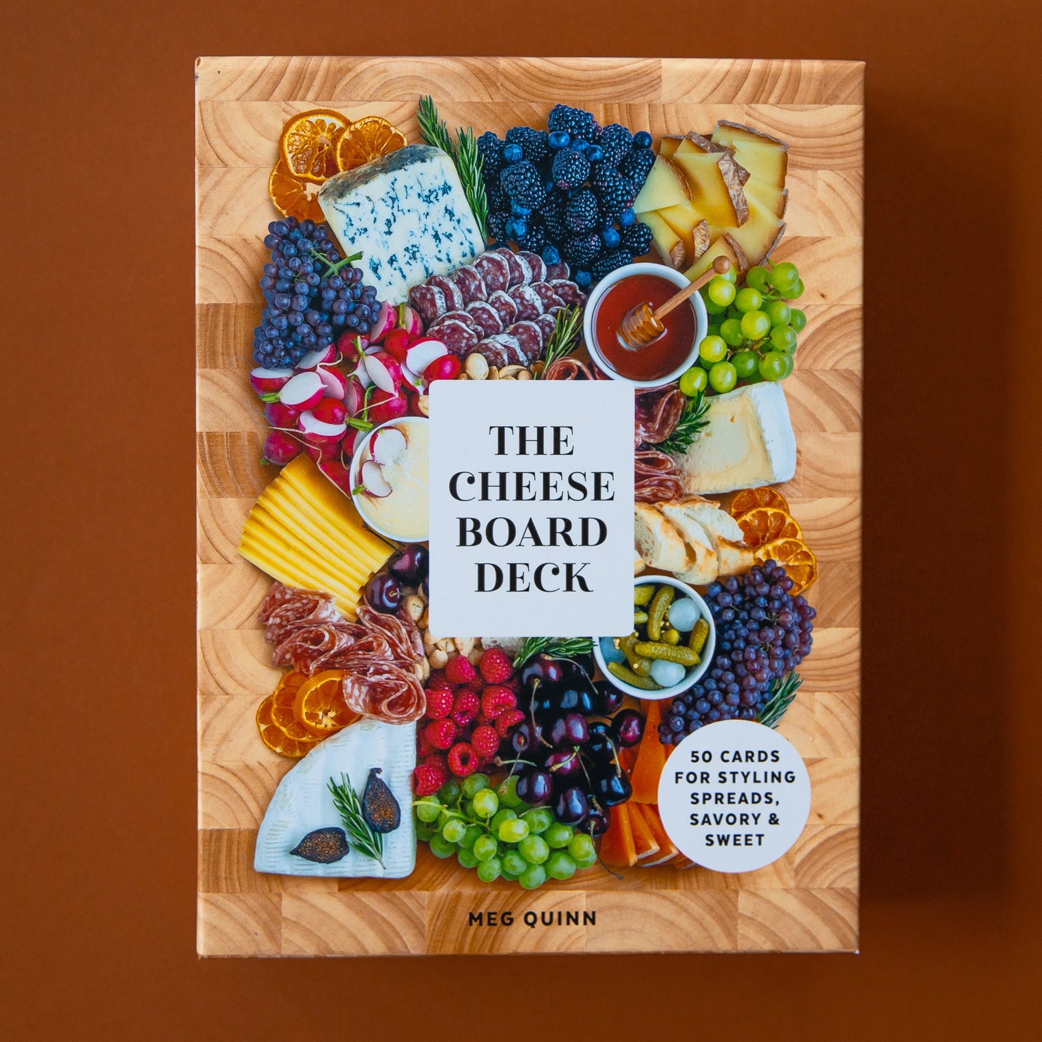 The front cover of box looks like a cheese board with an assortment of cheeses, meats and fruits along with a white square in the center that says, "The Cheese Board Deck".
