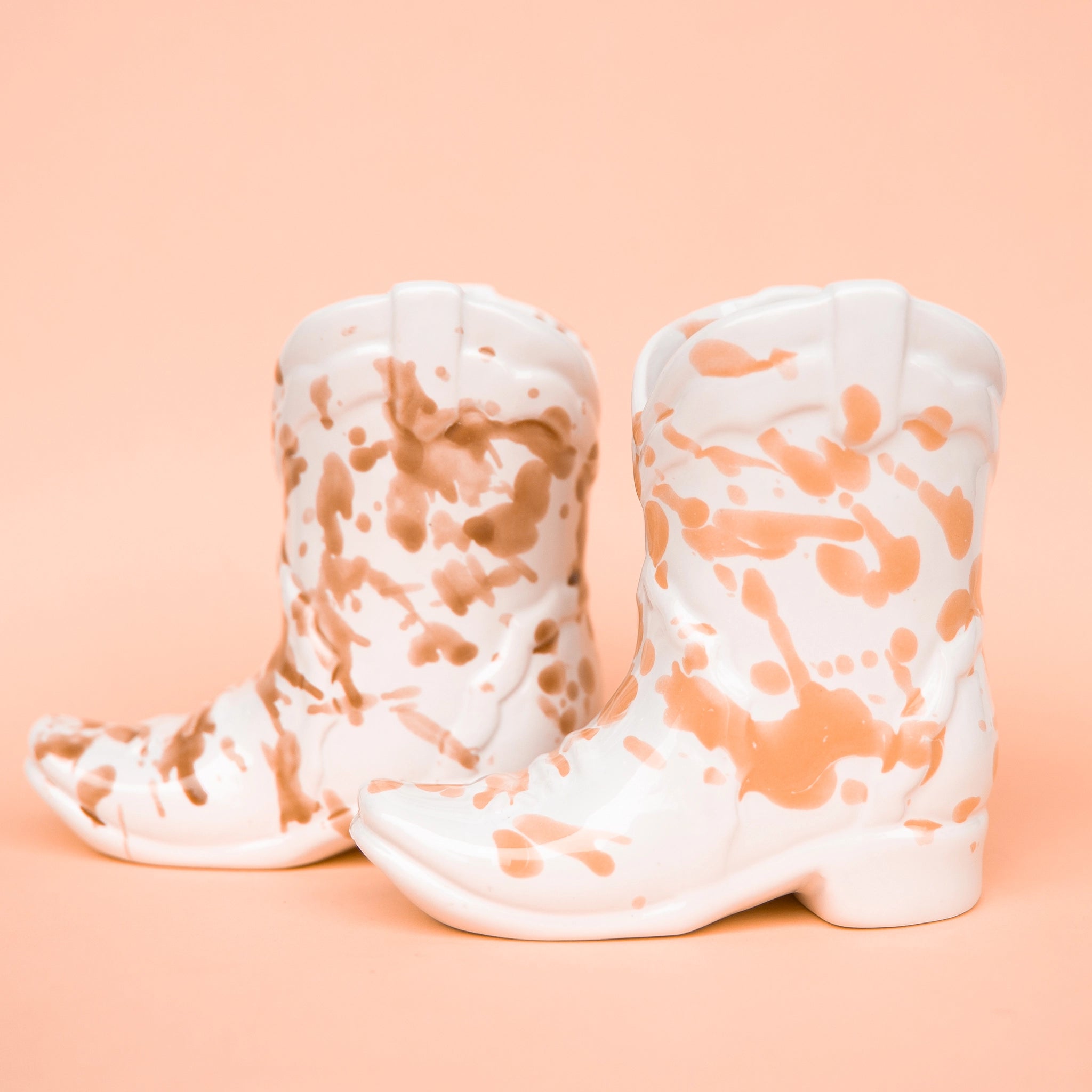 On a peachy background is two ceramic cowboy boot shaped candles with peachy and brown speckles. 