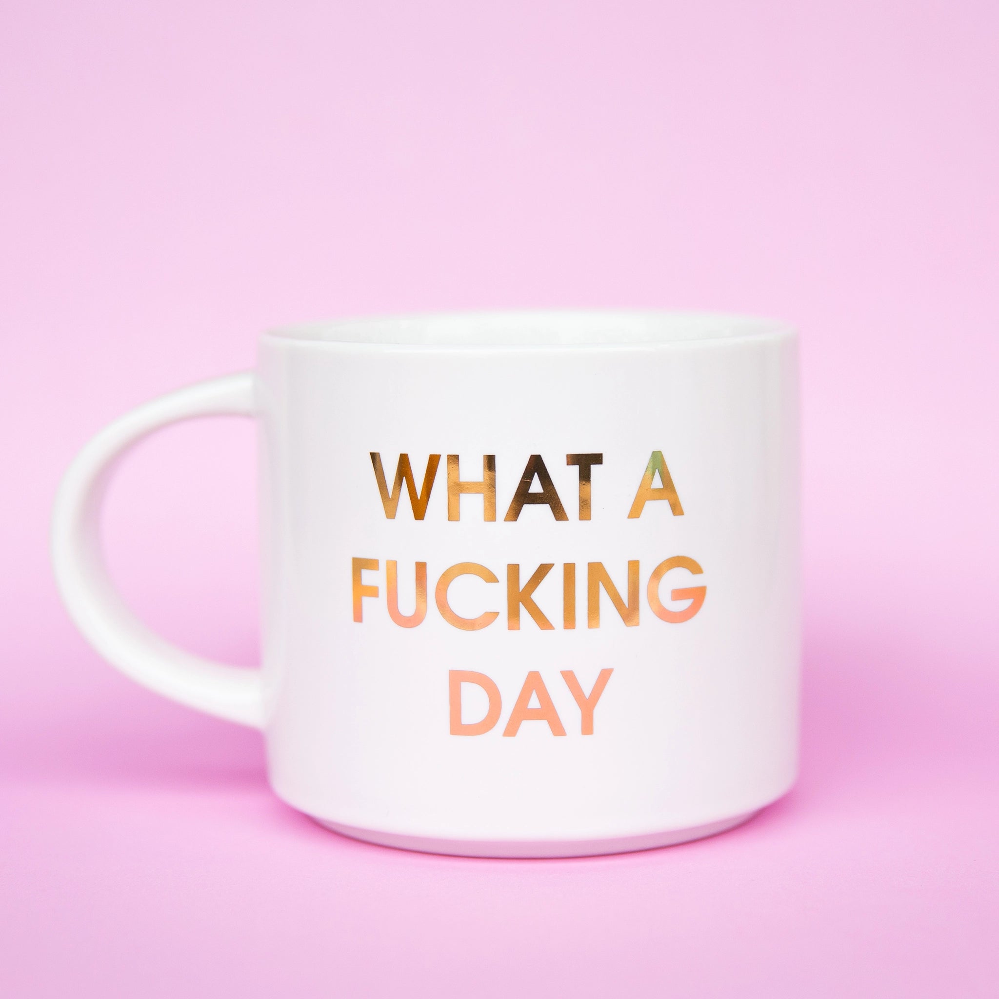Classic white ceramic mug labeled &#39;What a Fucking Day&#39; in gold shimmering capital lettering. Mug has a round white handle fasted on the left hand side.