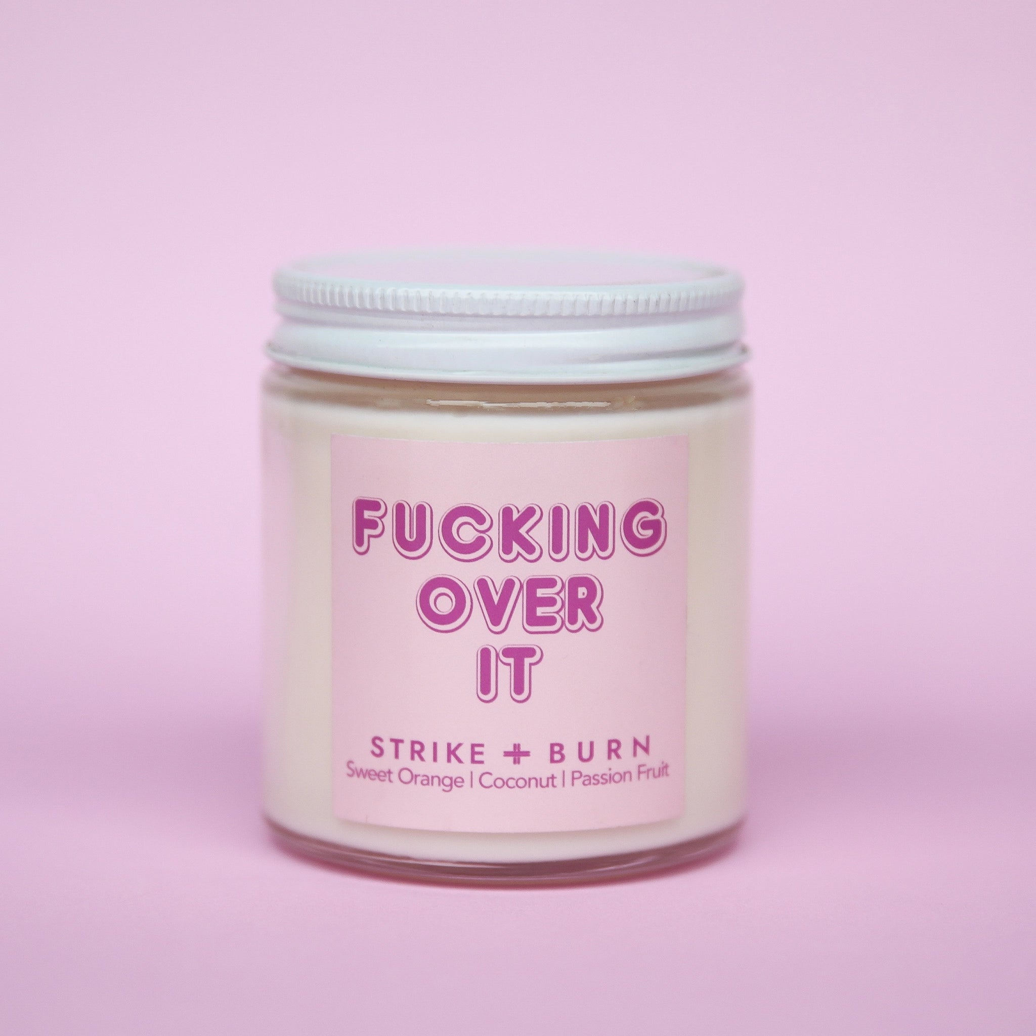 Clear glass jar candle with a white lid off to the side. The label is light pink with font that reads "fucking over it".