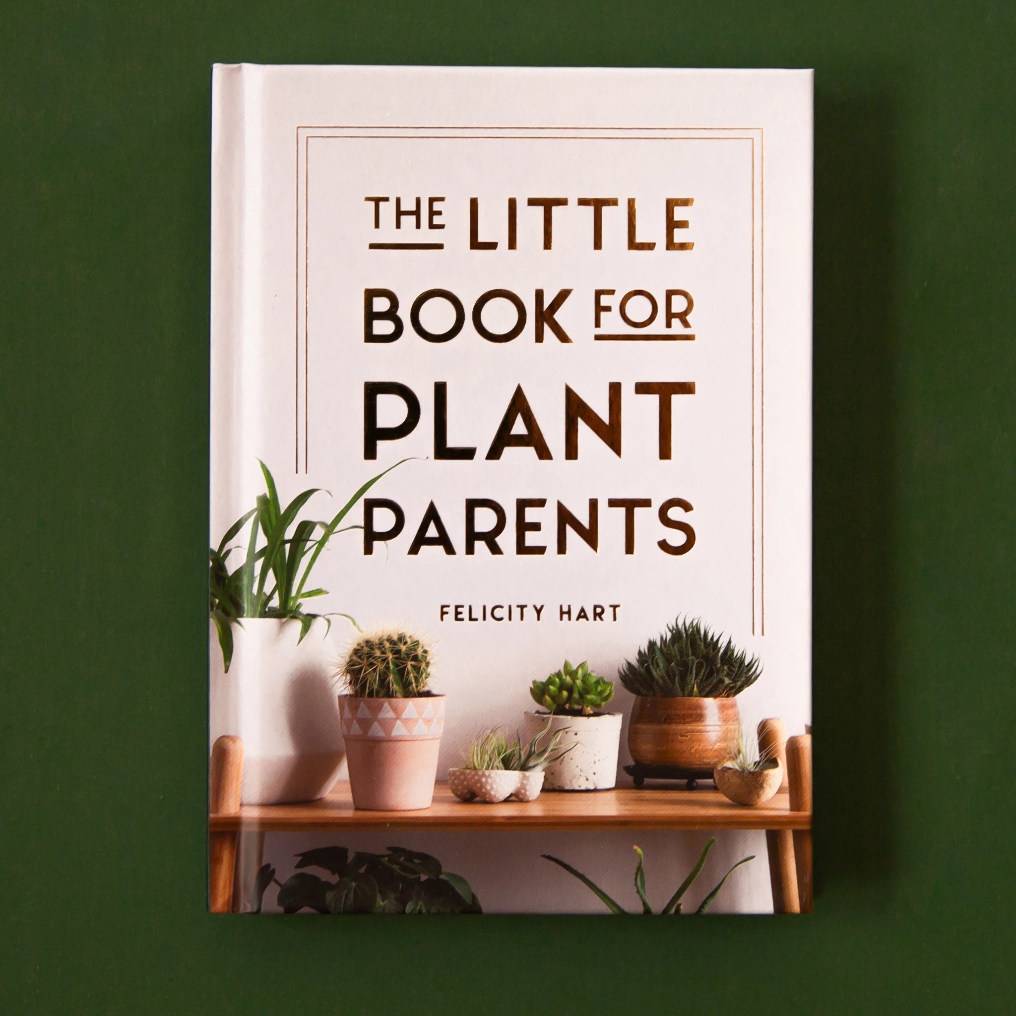On a cream background is a light tan/pink book cover with rose gold text that reads, "The Little Book For Plant Parents by Felicity Hart" as well as a photograph of different house plants and cacti sitting on a shelf.