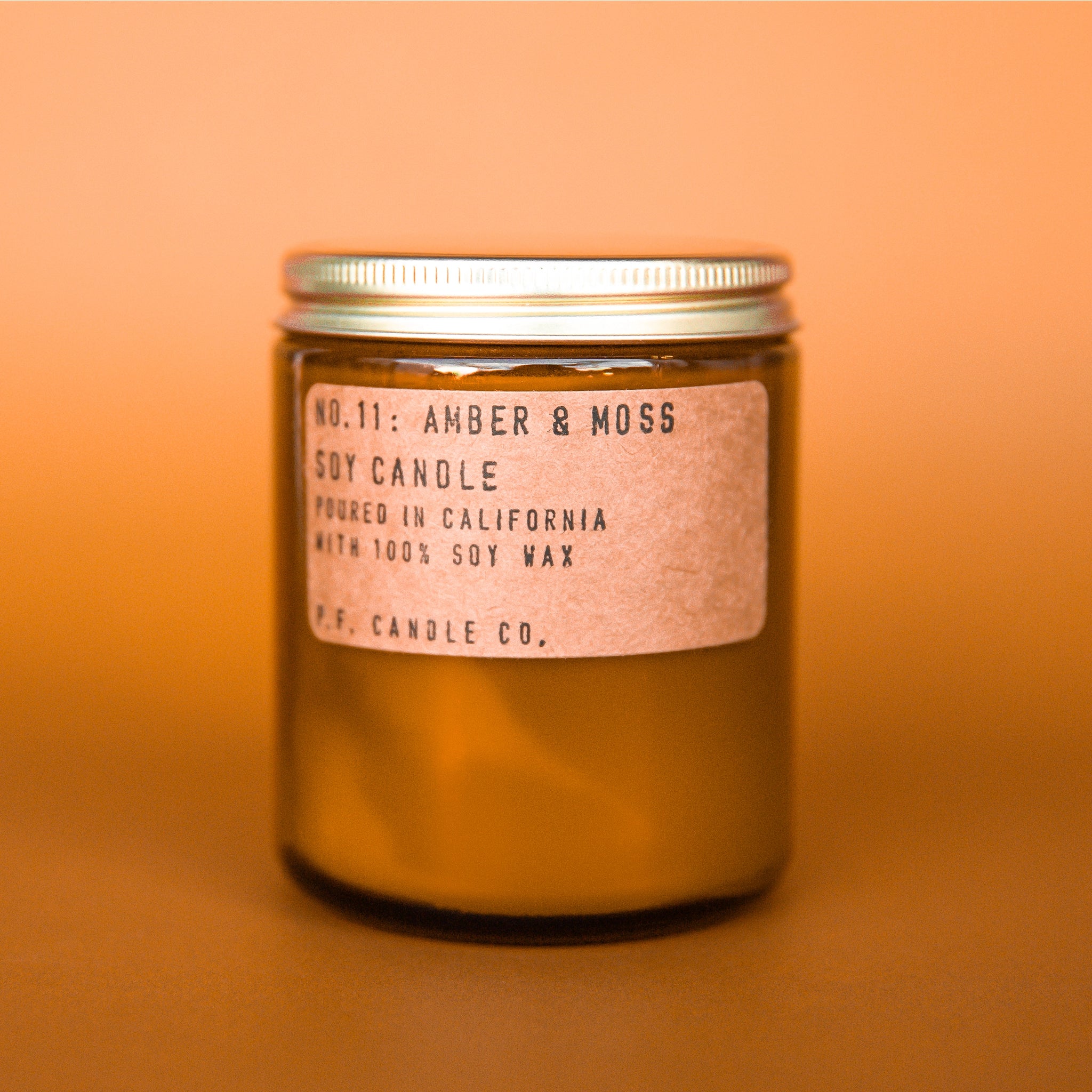 On an orange background is an amber glass jar candle with a kraft brown label and a gold lid. 
