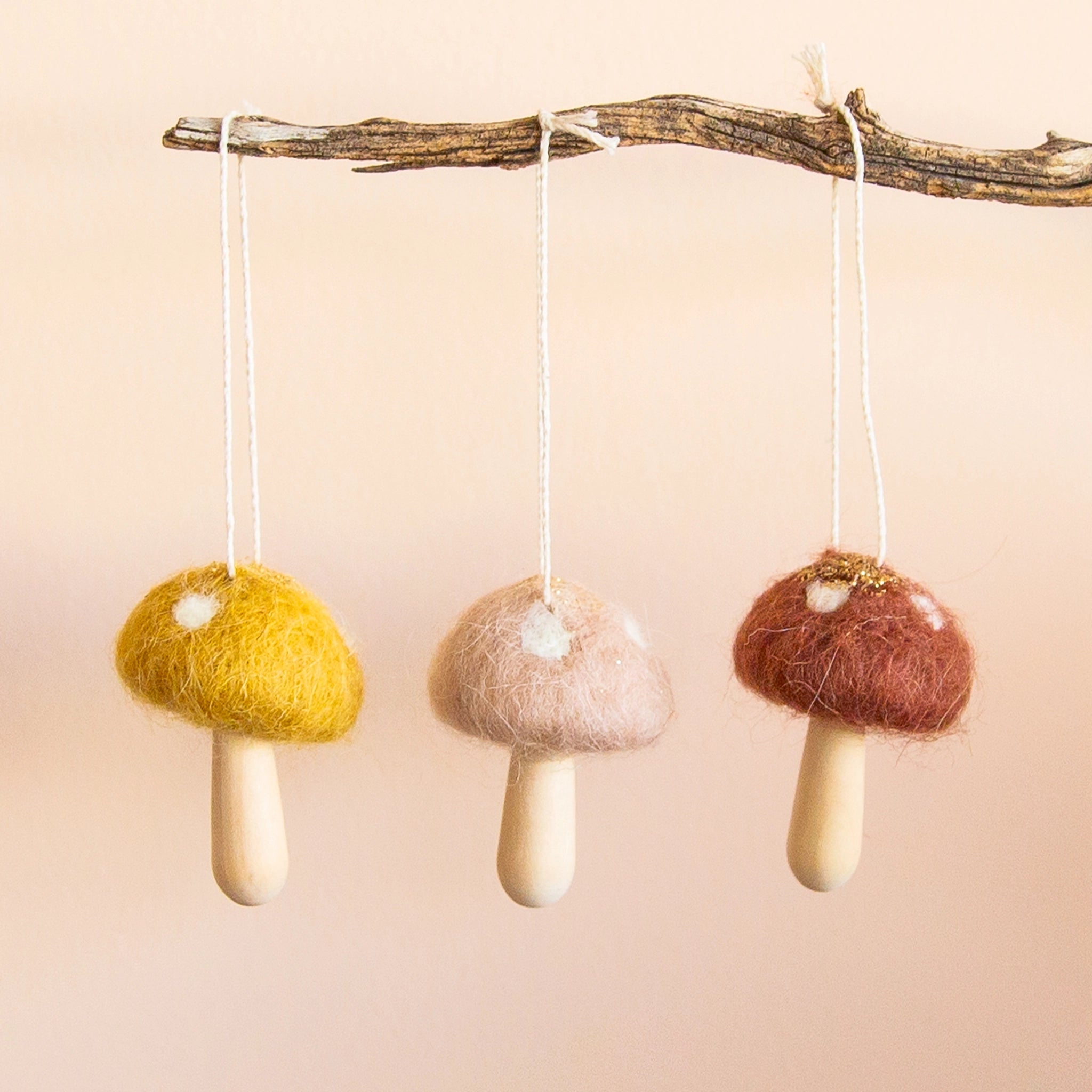 On a neutral background is three felt mushroom ornaments hanging on a stick. From left to right, there is a yellow mushroom, neutral tan and cocoa brown.