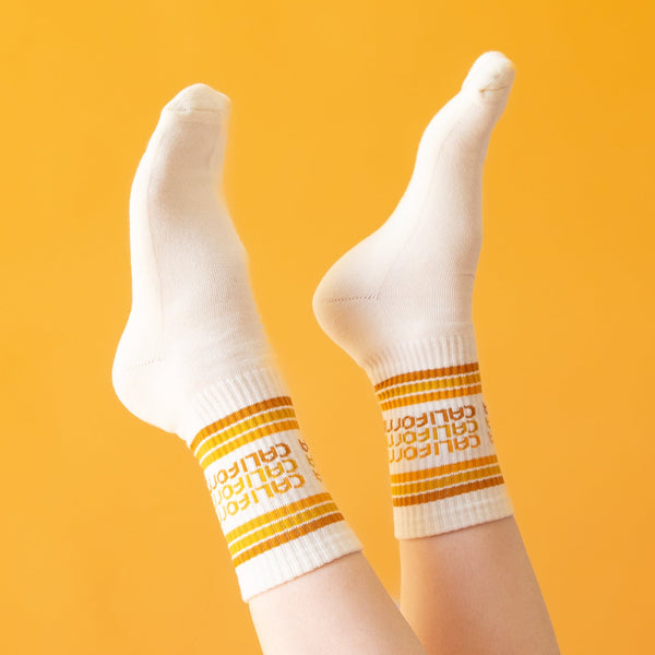 On a yellow background is a pair of white socks with rust, yellow and orange stripes and "california" printed three times on the ankle.