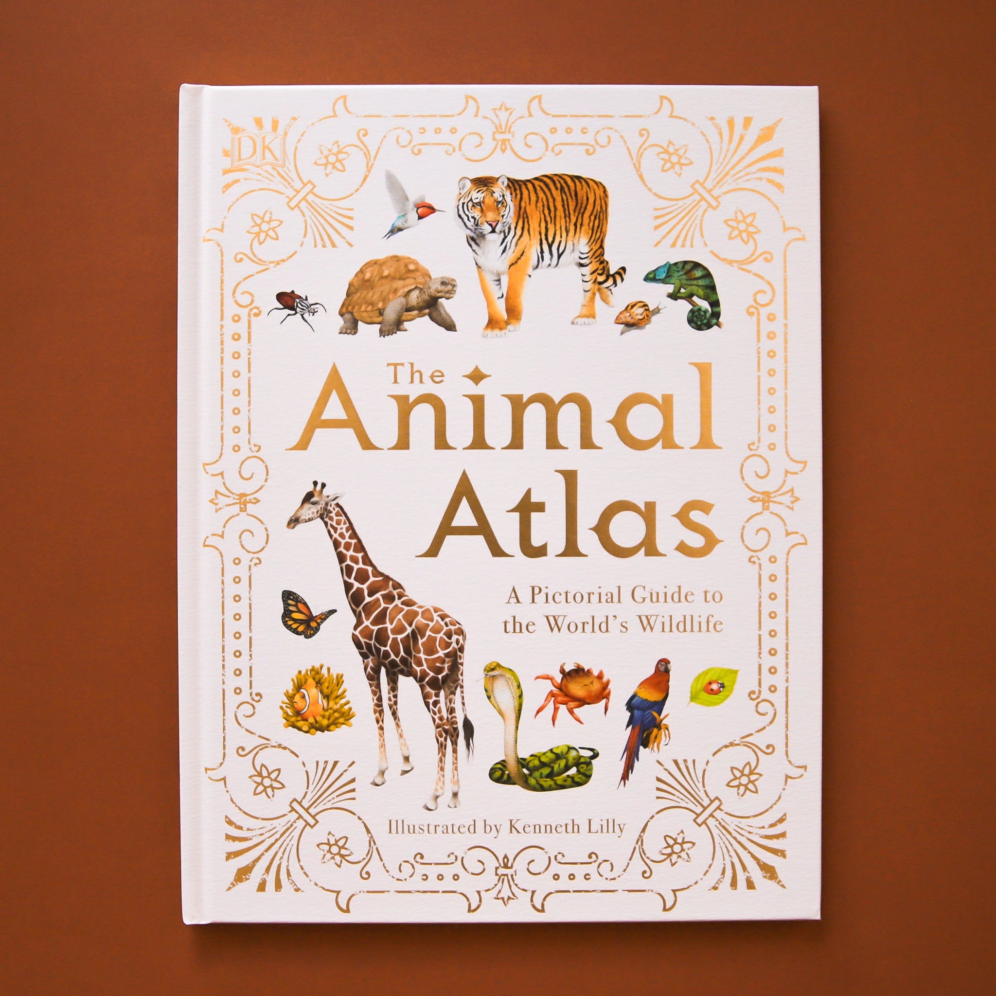 The front cover of the book reads &#39;The Animal Atlas&#39;: A pictorial Guide to the World&#39;s Wildlife&#39; in gold lettering. The text is surround by a variety of animals including tigers, turtles, giraffes, and more. The cover of the book is bordered with elegant gold detailing.