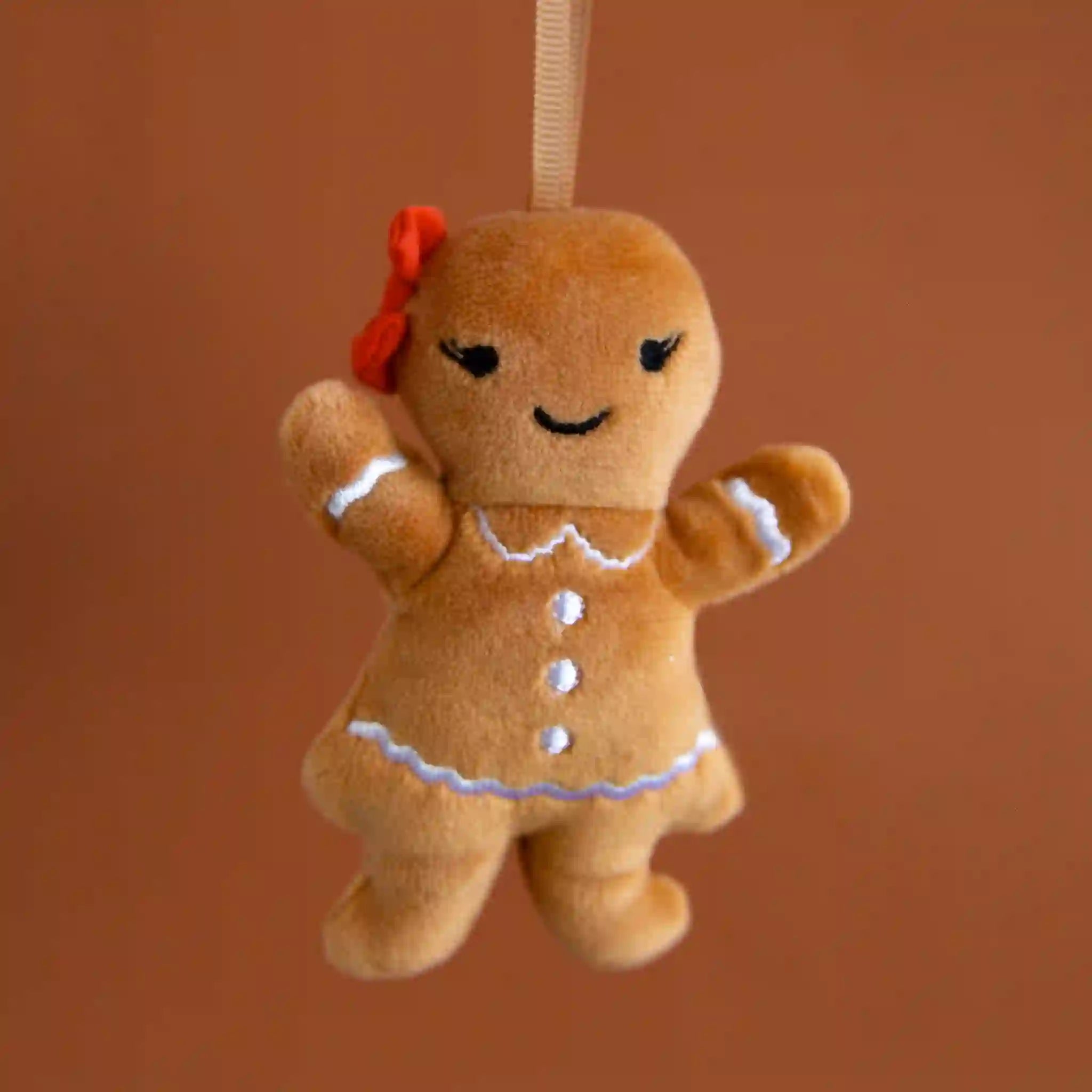On a brown background is a brown gingerbread shaped stuffed toy with a smiling face, white detailing and a red bow on her head.
