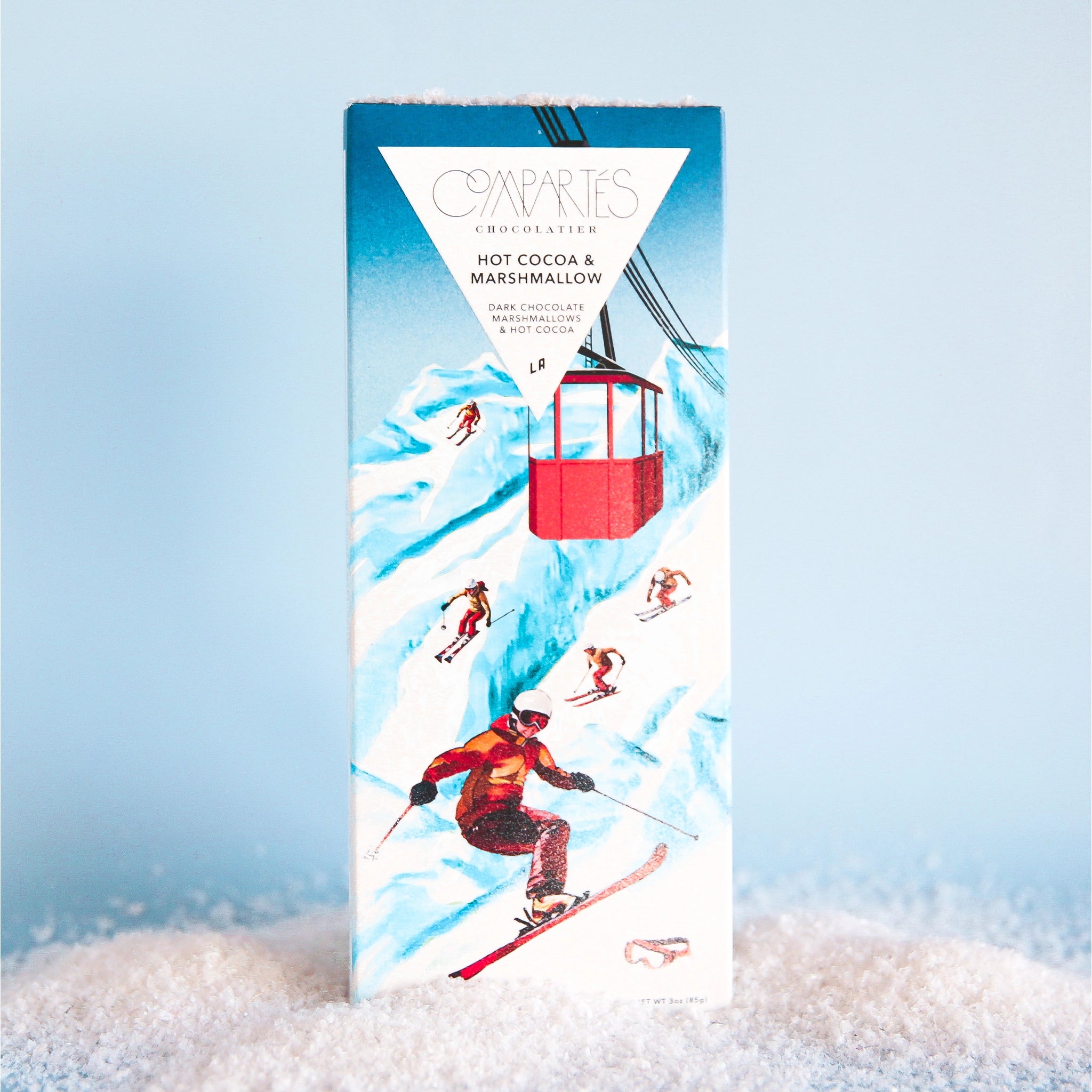 On a light blue background is a rectangle chocolate bar with packaging that features a mountain and skiers in red outfits along with text at the top that reads, "Compartes Hot Cocoa & Marshmallow Dark Chocolate".
