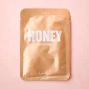 On a pink background is a gold packet holding a face mask with white text across the front that reads, "Honey Nourishing". 