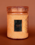 On a burnt orange background is a large decorative jarred candle in an orange shade with a matching lid and black label in the center that reads, "Voluspa Spiced Pumpkin Latte".