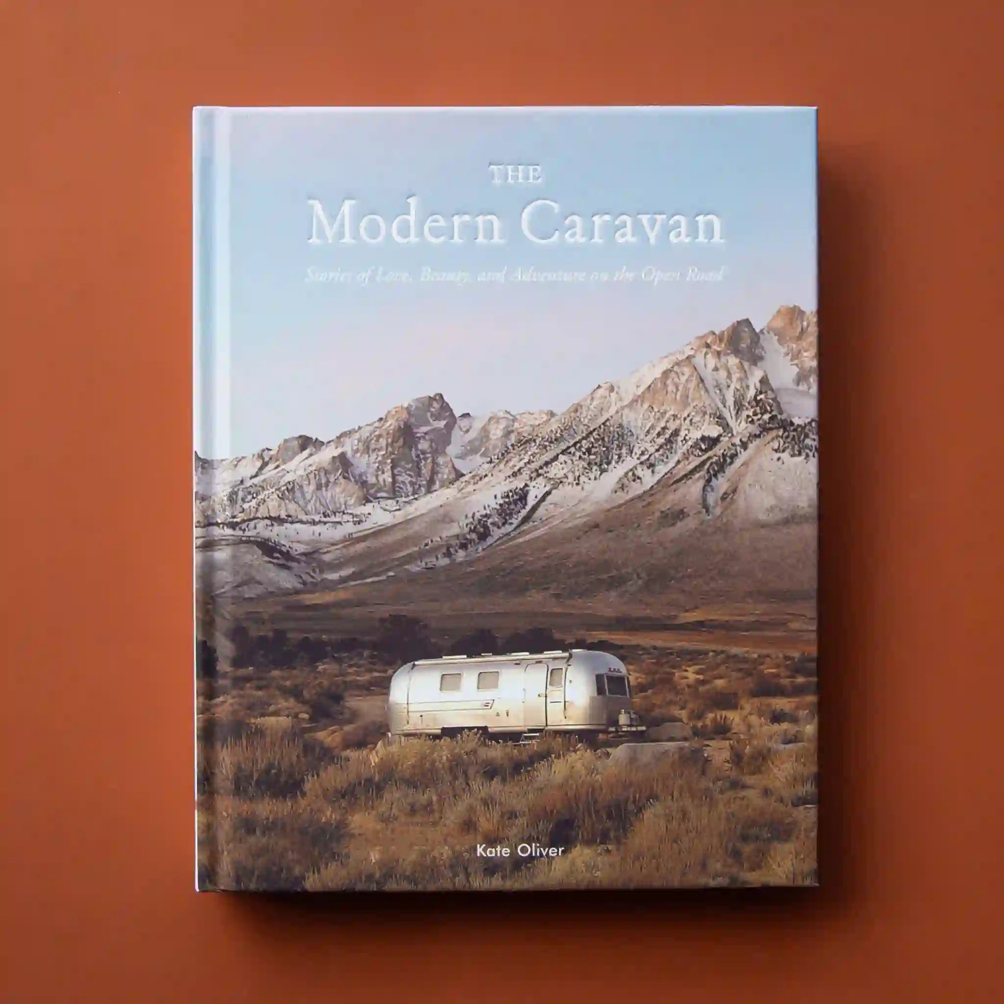 Hard cover of travel book titled 'The Modern Caravan, stories of love, beauty, and adventure on the open road' in white pressed lettering. Behind the title is a cool toned mountain scene. Below the mountains sits a silver airstream trailer amidst an open valley.