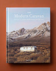 Hard cover of travel book titled 'The Modern Caravan, stories of love, beauty, and adventure on the open road' in white pressed lettering. Behind the title is a cool toned mountain scene. Below the mountains sits a silver airstream trailer amidst an open valley.