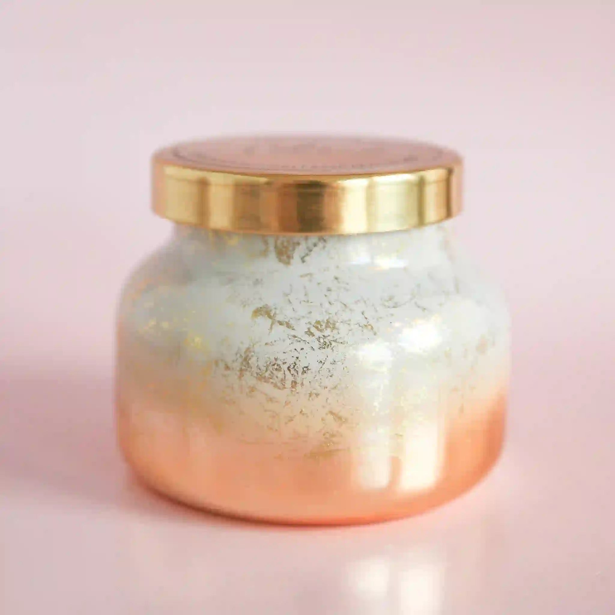 On a pink background is a white and gold glass jarred candle with a gold lid that reads, "capriBLUE Volcano"