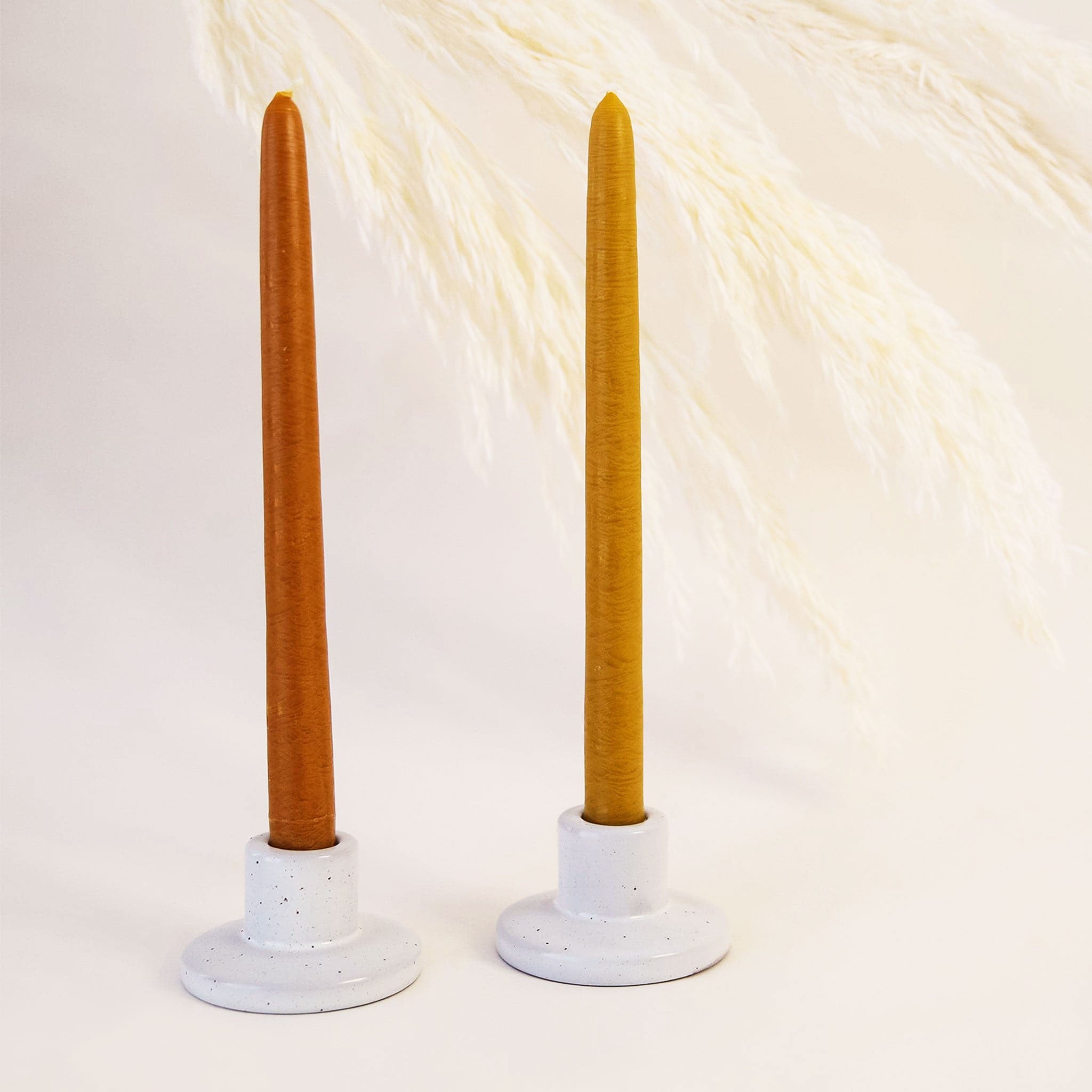 In front of a white background is two ceramic candle holders. The gray speckled candle holders have a round base with a short tapered top. Inside the right candle holder is a mustard yellow candle stick and in the left is a dark orange candle stick.