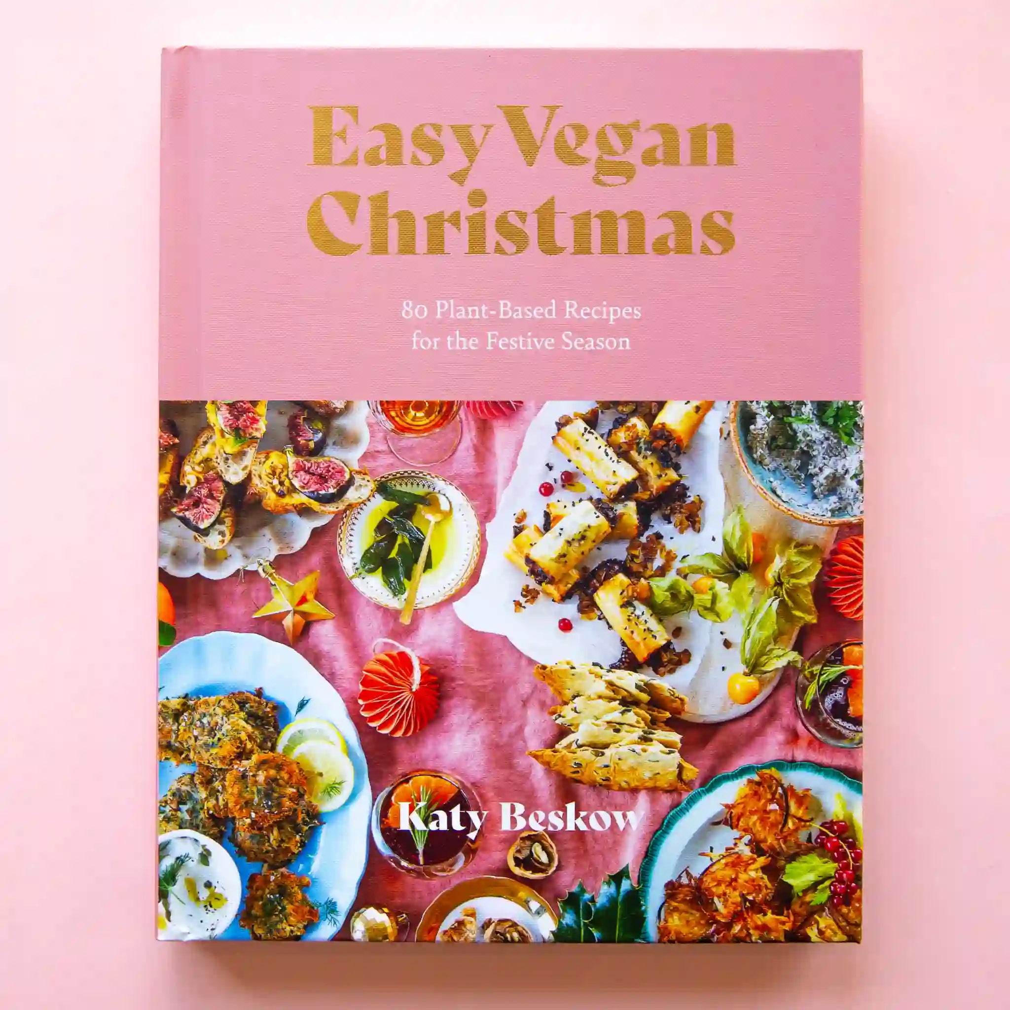 On a pink background is a pink and colorful book cover with gold text on the top that reads, "Easy Vegan Christmas", "80 Plant-Based Recipes for the Festive Season" along with a photo of colorful food laid out on a table scape. 