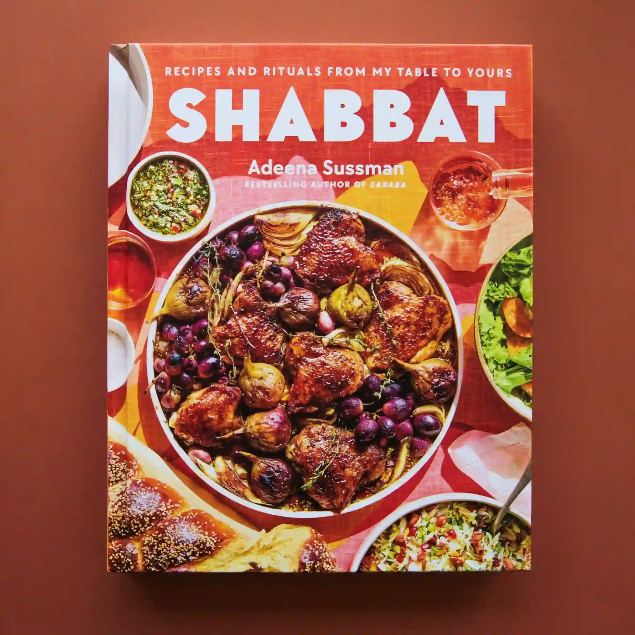 On a burnt orange background is a colorful book cover with a big family style meal with white text at the top that reads, &quot;Recipes and RItuals From My Table To Yours Shabbat&quot;.