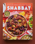 On a burnt orange background is a colorful book cover with a big family style meal with white text at the top that reads, "Recipes and RItuals From My Table To Yours Shabbat".