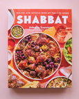 On a pink background is a colorful book cover with a big family style meal with white text at the top that reads, "Recipes and RItuals From My Table To Yours Shabbat".