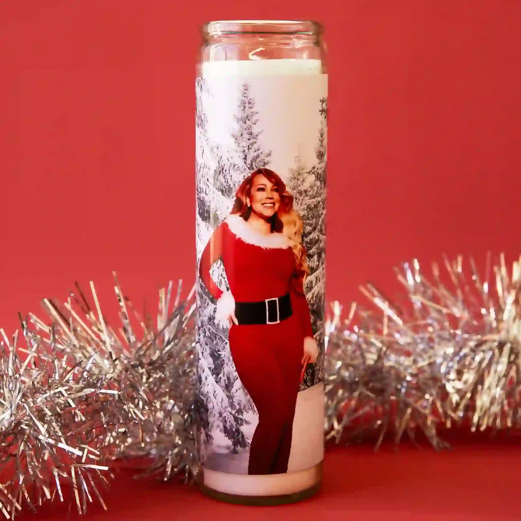 On a red background is a thin glass prayer candle with a graphic of Mariah Carey in front of a white snowy background wearing a red Mrs. Clause dress. 