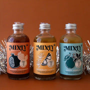 On a brown background is a modern cocktail kit with three glass bottles of cocktail mixes.