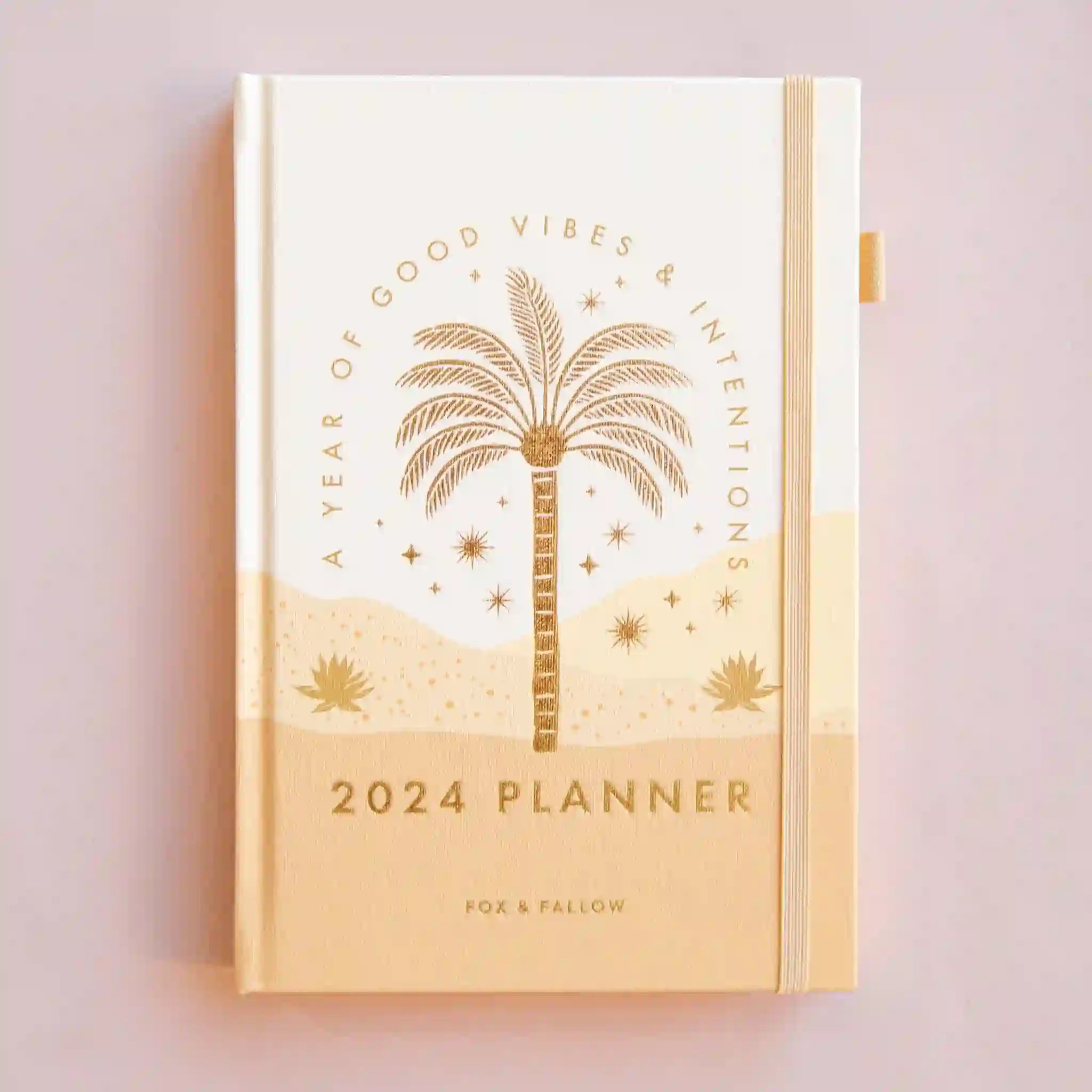 On a pink background is a tan planner with gold foiled "2024 Planner" text in the center as well as arched text over a palm tree design that reads, "A Year Of Good Vibes & Intentions" and a light pink elastic loop for keeping shut.