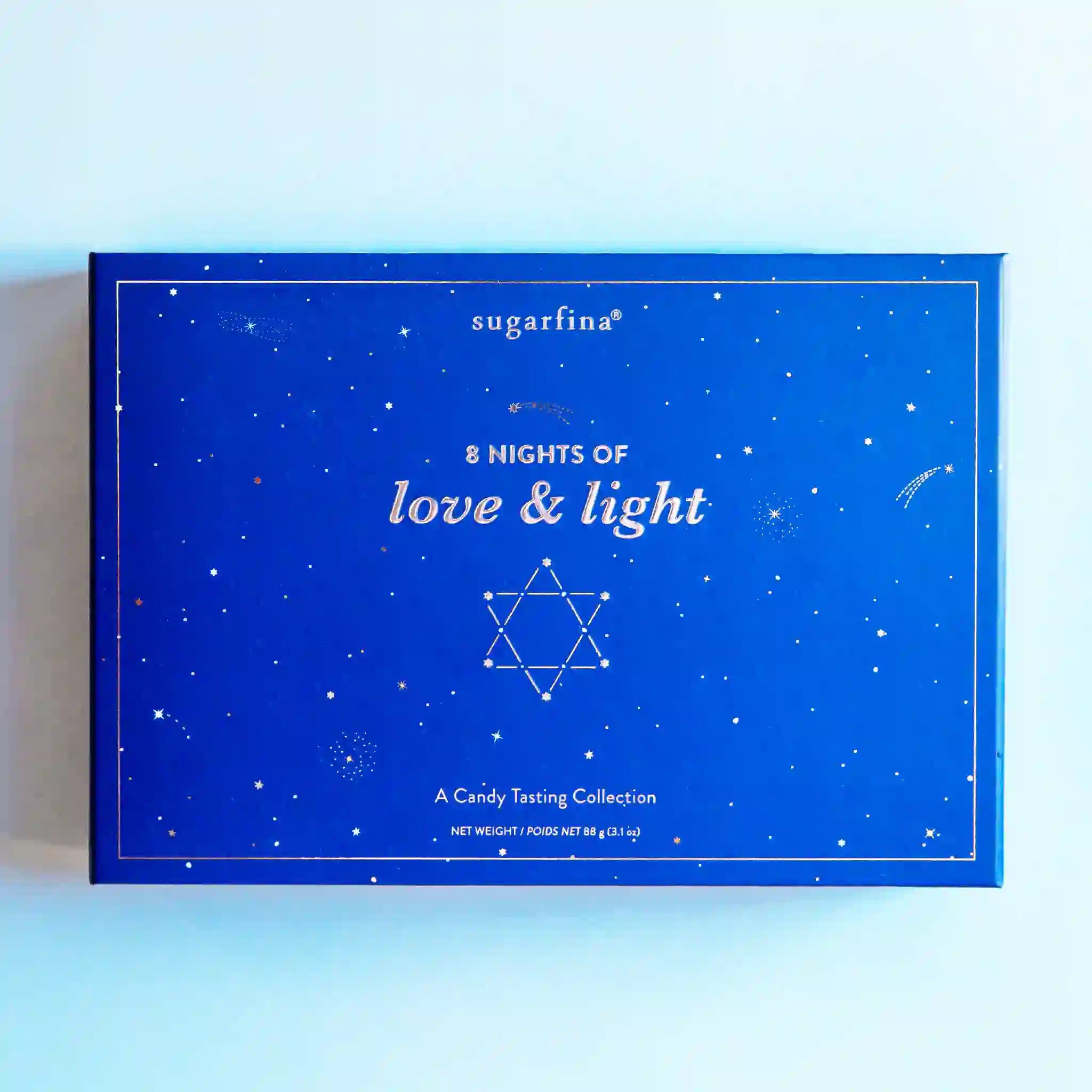On a light blue background is a blue box filled with an assortment of chocolate and candies for each night of Hanukkah. The front of the box features silver details and text that reads, "8 Nights of love & light A Candy Tasting Collection".