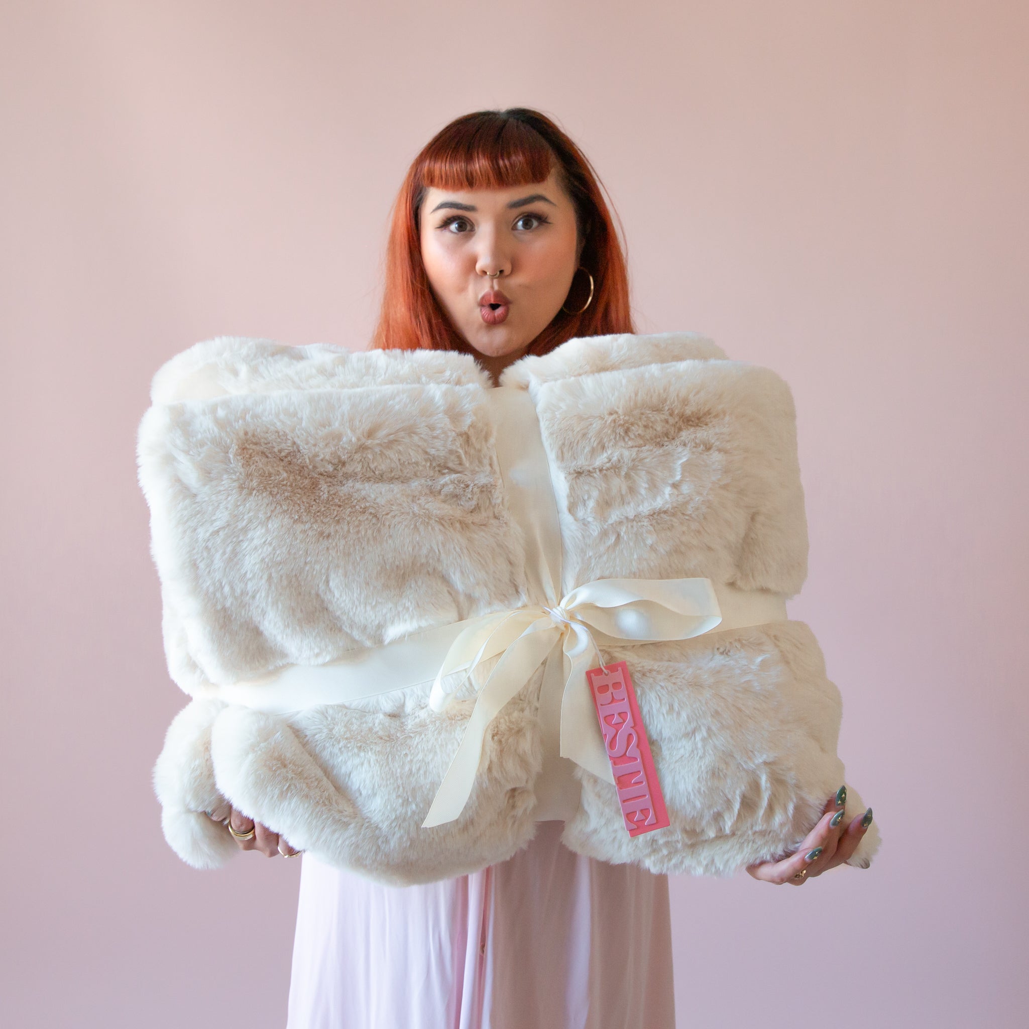 A red headed woman holds a giant white fur blanket in her hands and stands in front of a pink background.