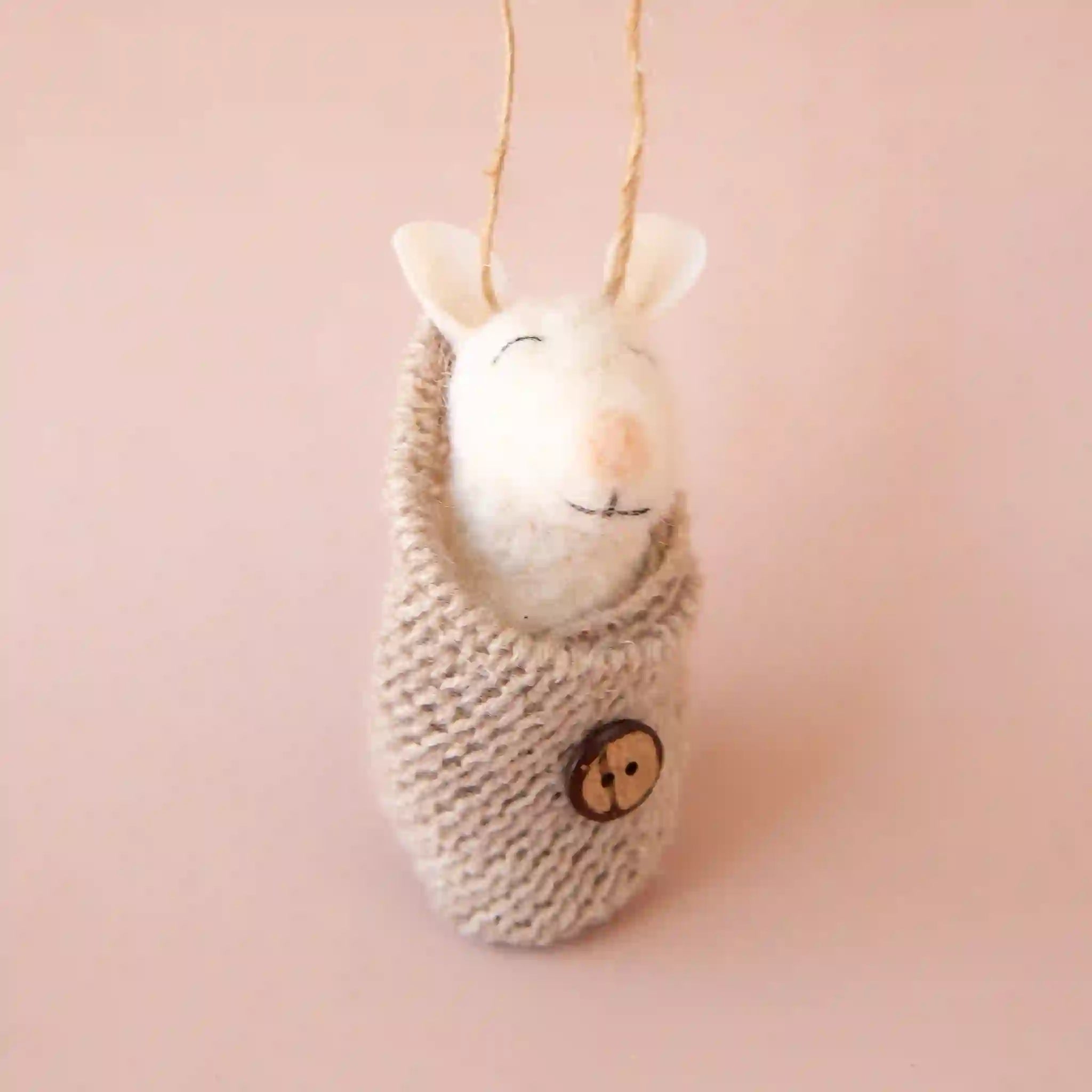 On a pink background is a felt baby mouse shaped ornament wrapped in a tan knit swaddle fastened with a button.