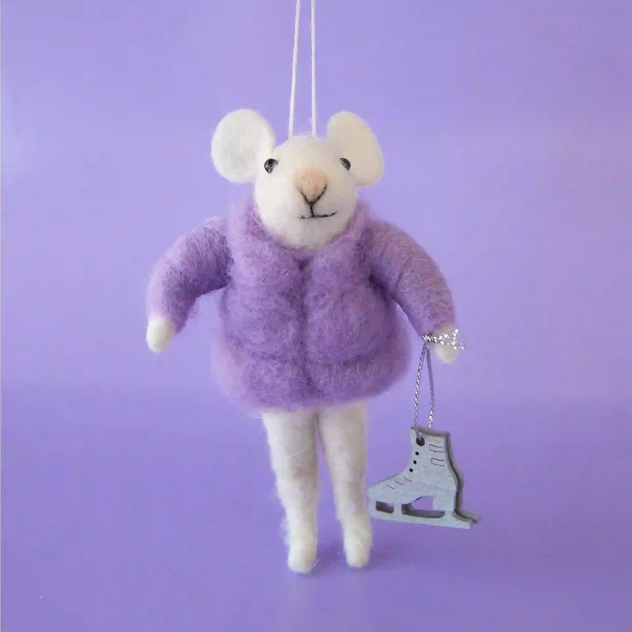 On a purple background is a white felt mouse ornament wearing a  purple puffy jacket and holding ice skates. 