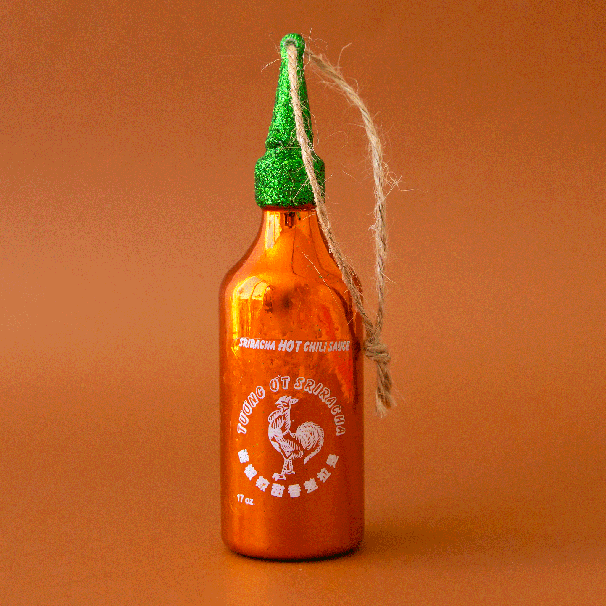 On a orange background is a red and green glass sriracha bottle shaped ornament with the classic logo int he center and a twine loop for hanging.