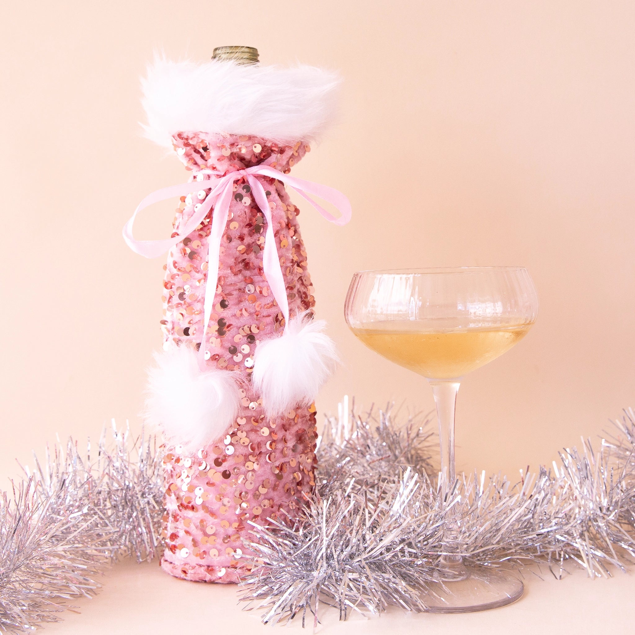 On a peach background is a sequin wine bag alongside a coupe glass. 