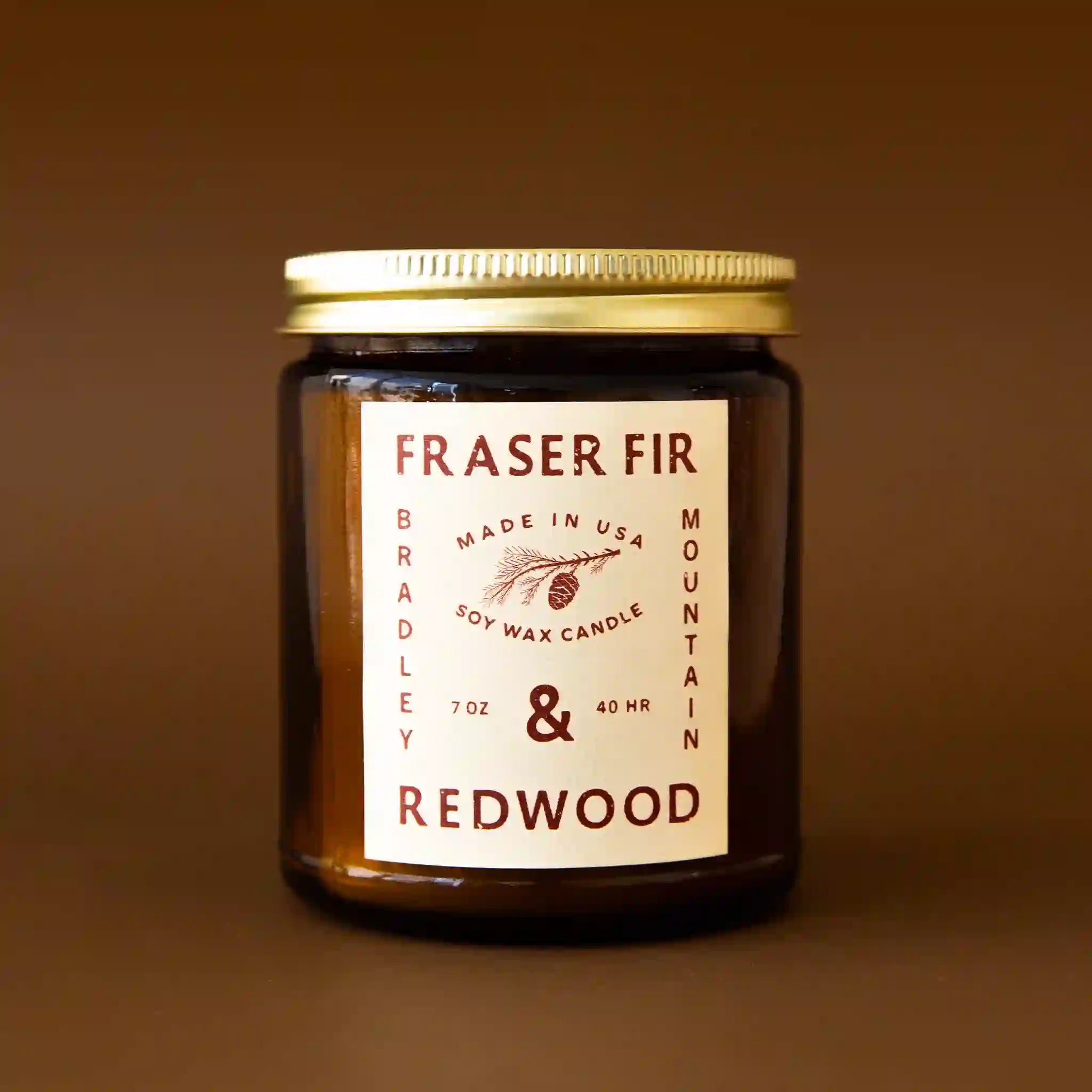 On a brown background is an amber brown glass candle jar with a tan label that reads, "Fraser Fir & Redwood".