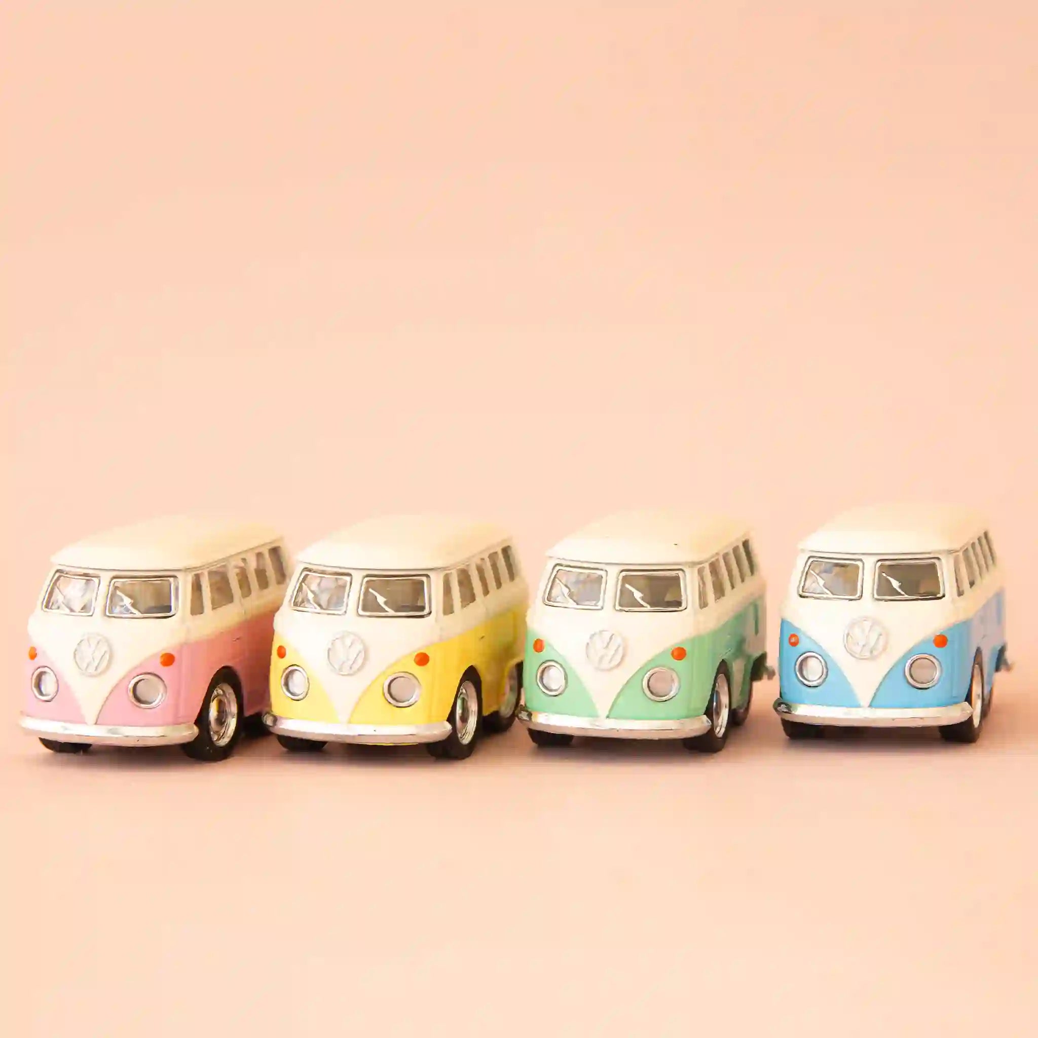 On a peach background are four VW toy buses in different colors. From left to right there is pink, yellow, green and blue. 
