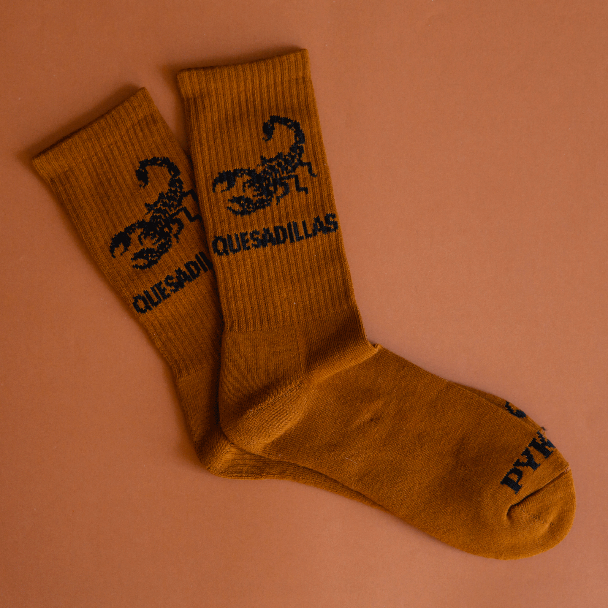 A pair of burnt orange crew socks with a black scorpion graphic and text that reads, "Quesadilla".