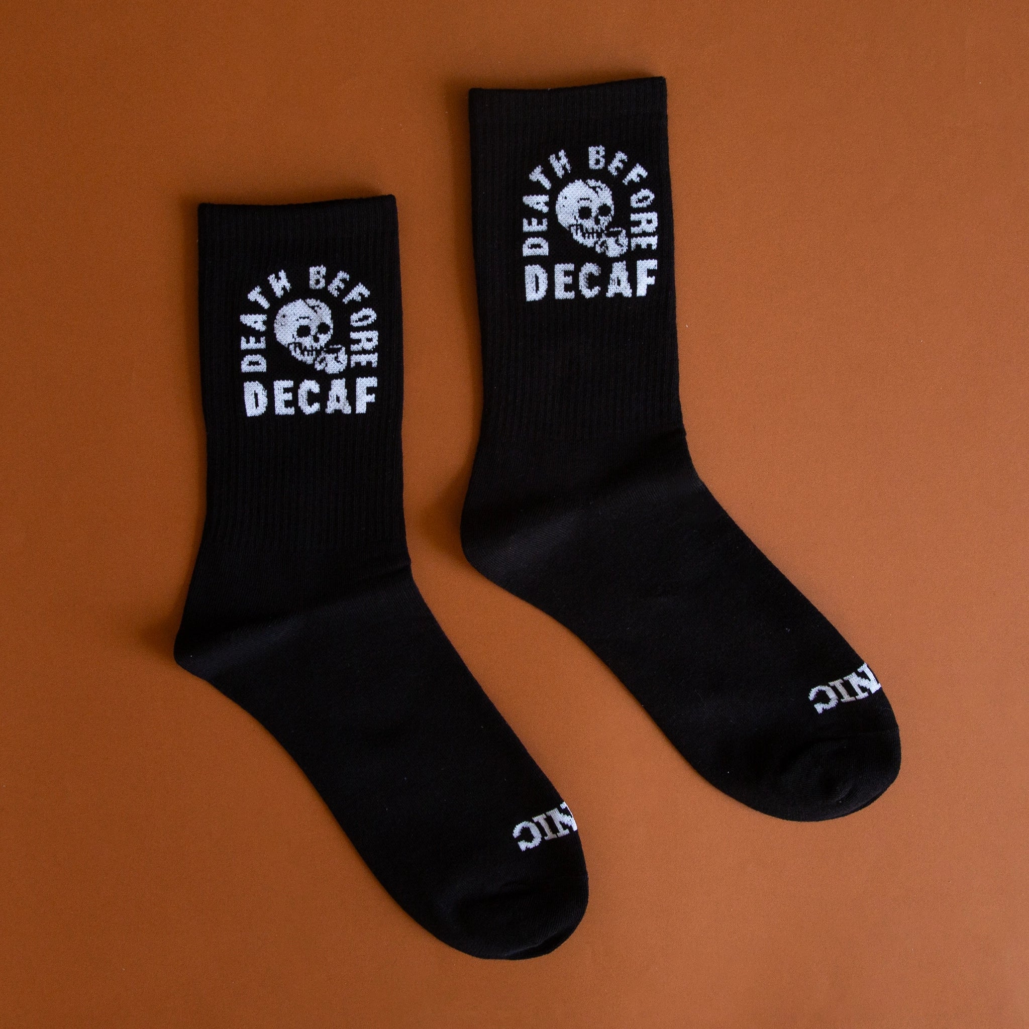 Black crew socks with a skull graphic on the ankle area and has white text wrapped around it that says, &quot;Death Before Decaf&quot;.