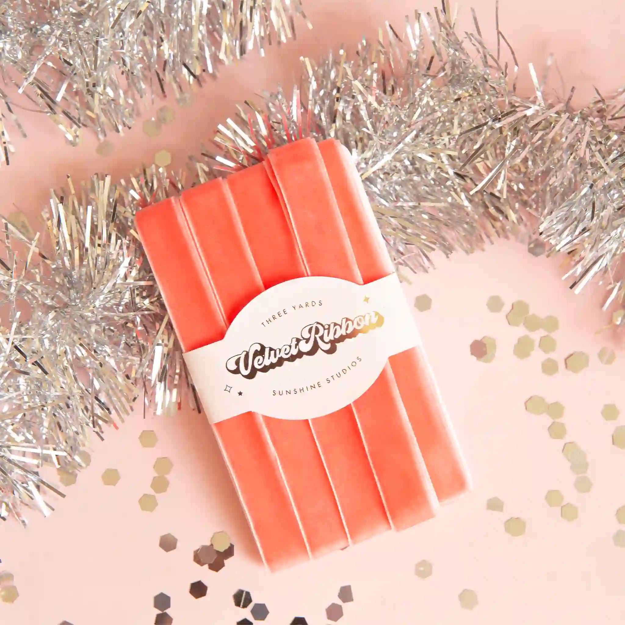 On a pink and silver tinsel background is a peach colored spool of velvet ribbon. 