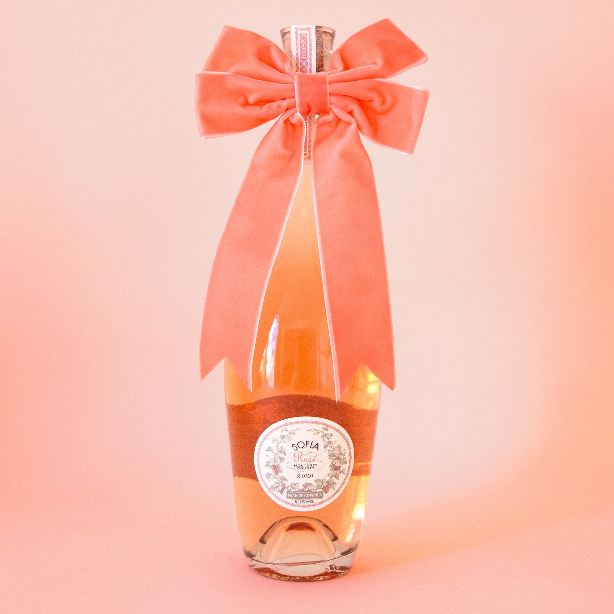 On a peachy background is a bottle of wine with a velvet peach colored bow on.