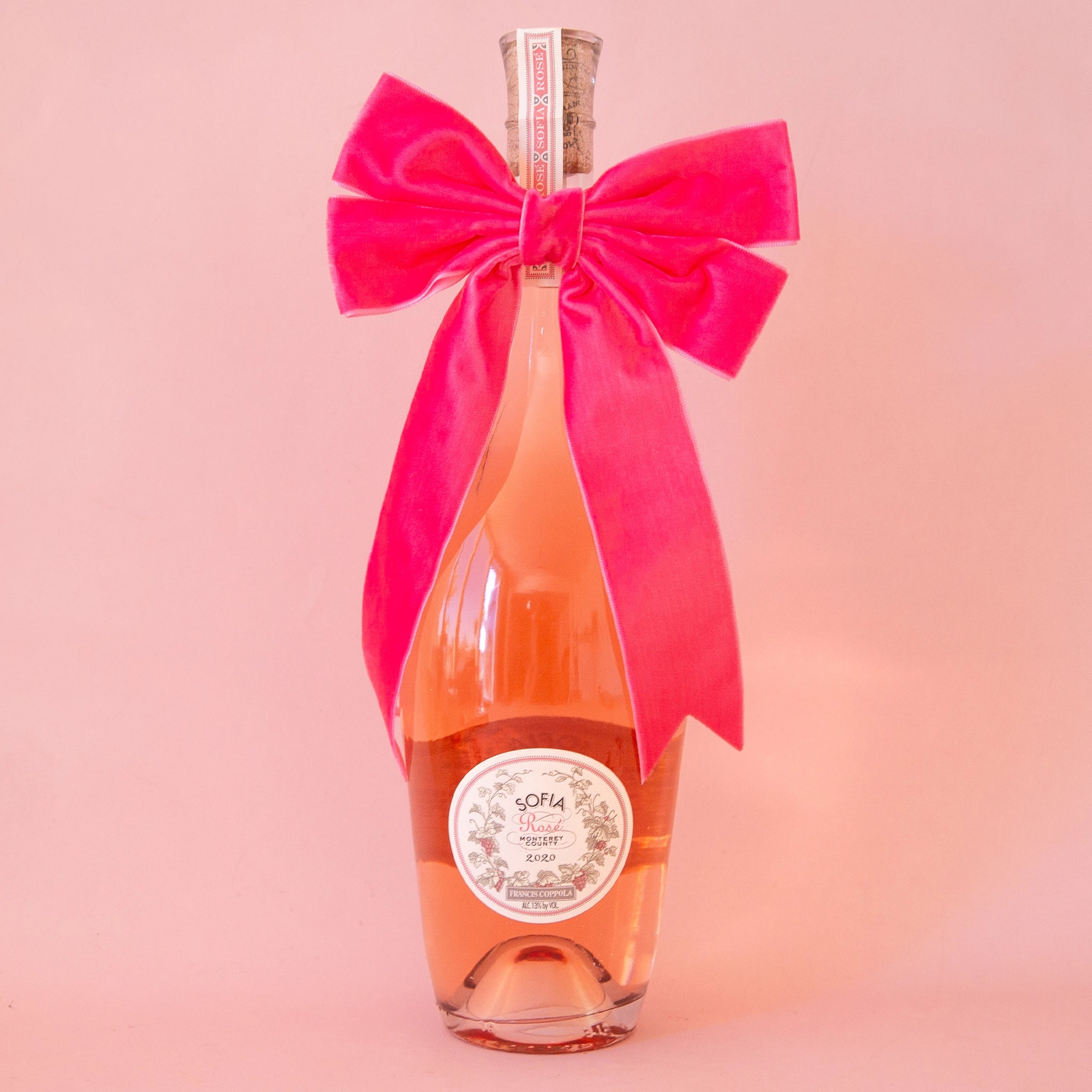 On a peachy background is a wine bottle with a large velvet pink bow
