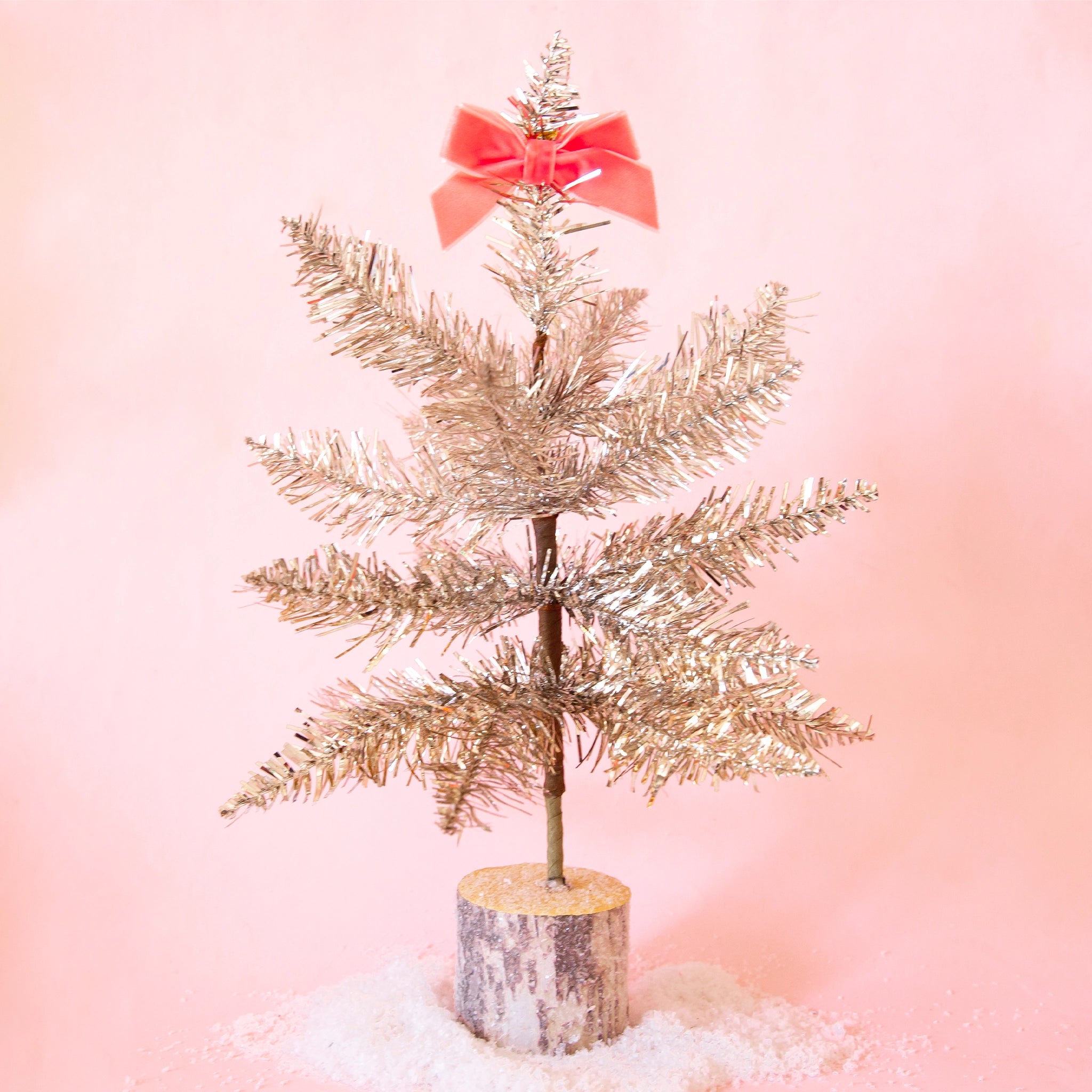 On a pink background is a silver tinsel tree with a wood base and a pink bow on top that is not included with purchase. 