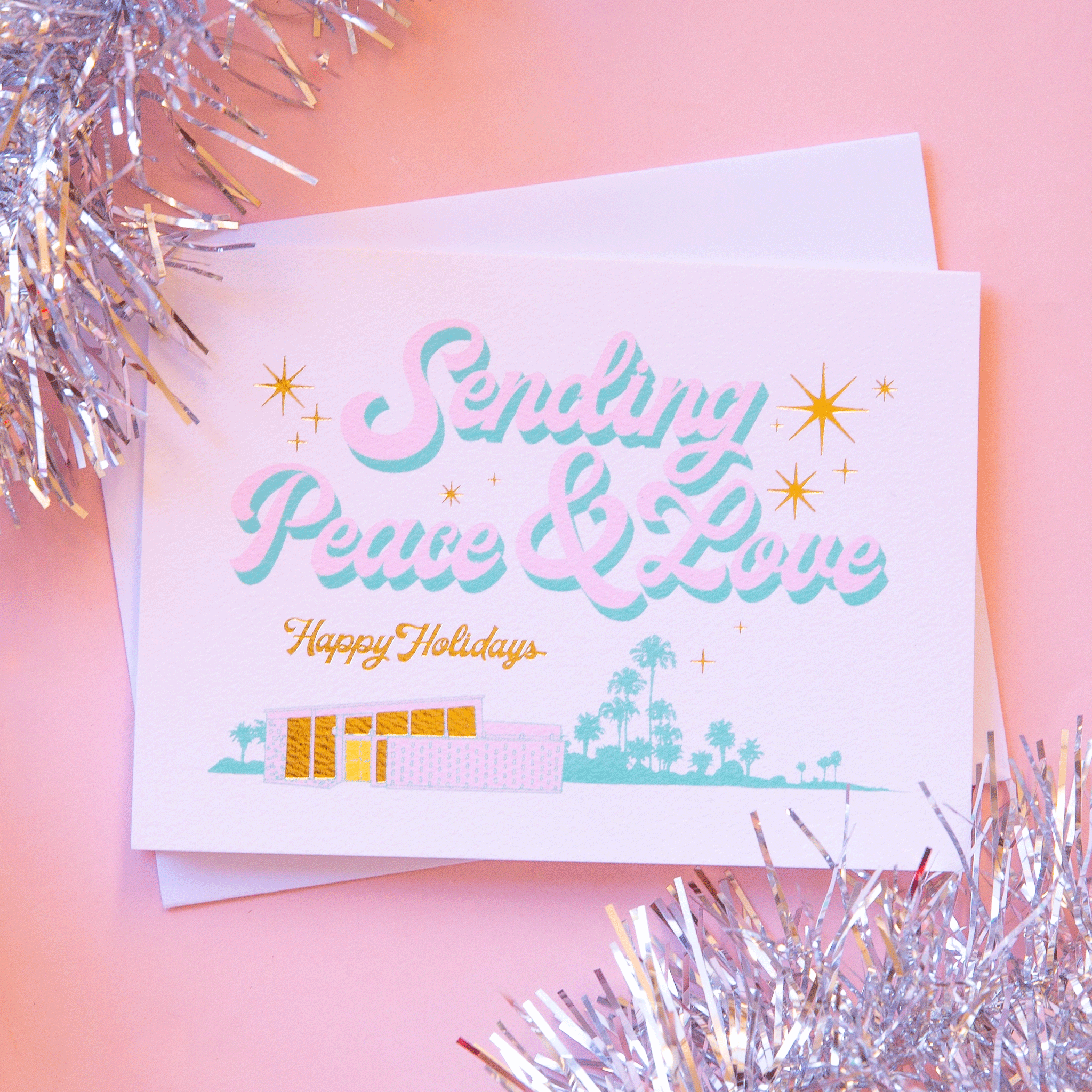 On a pink background is a white, pink and blue holiday card that reads, "Sending Peace & Love Happy Holidays".