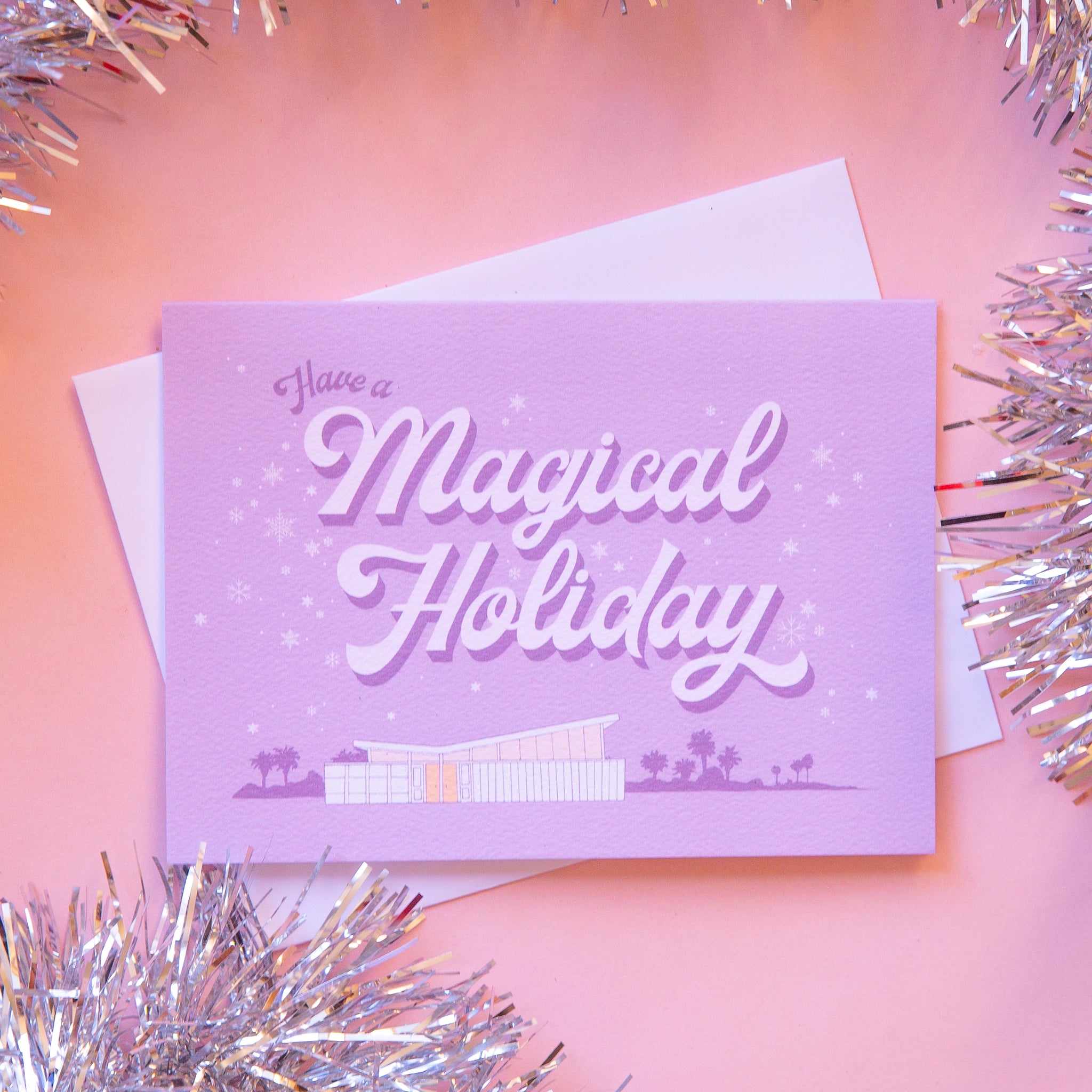 Lilac holiday card that reads &#39;have a a magical holiday&#39; in cursive lettering. Around the text is delicate white snowflakes. Below is a coastal scene of a beach house and palm trees.