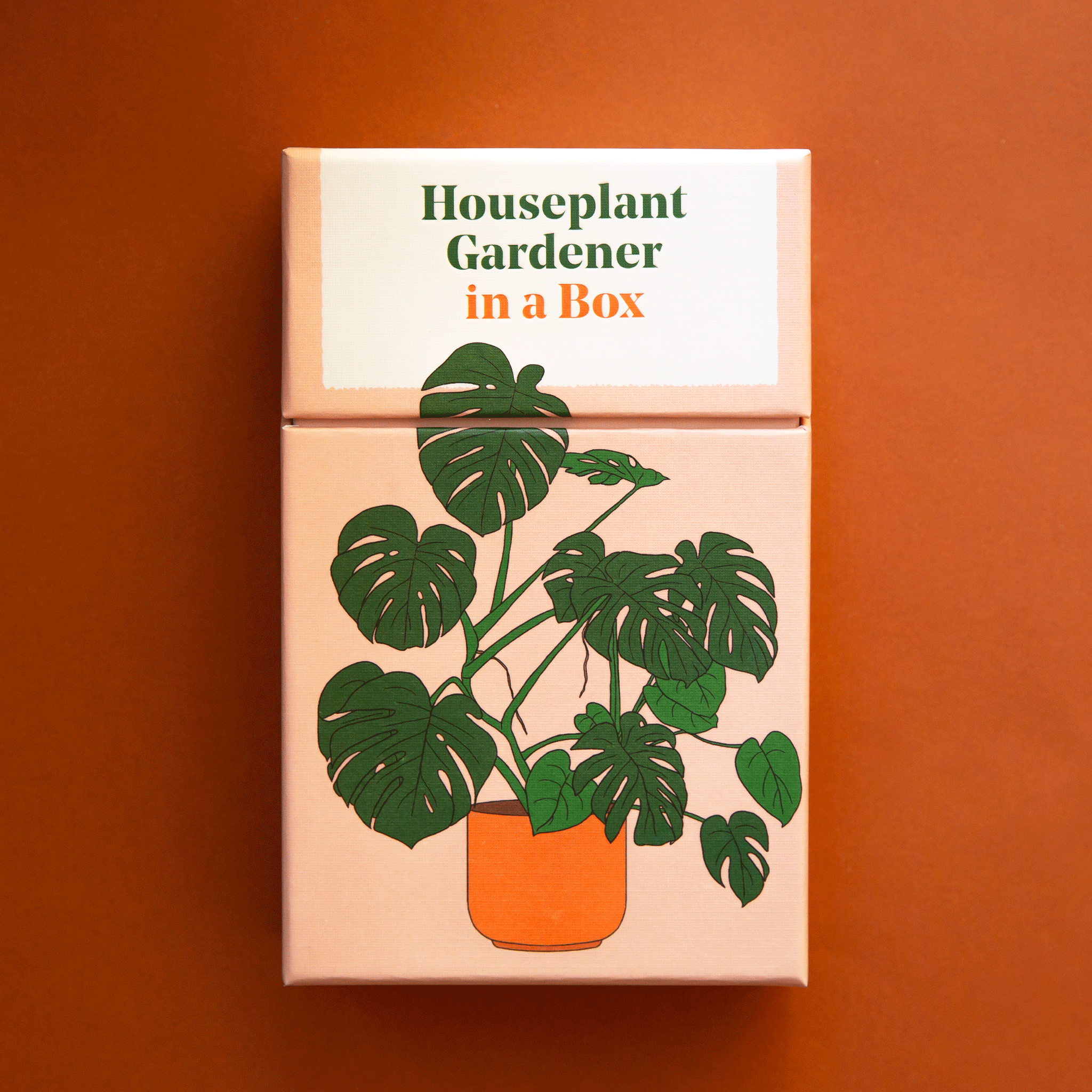 On an orange background is a tan box with an illustration of a split leaf with text that reads, "Houseplant Gardener in a Box". 