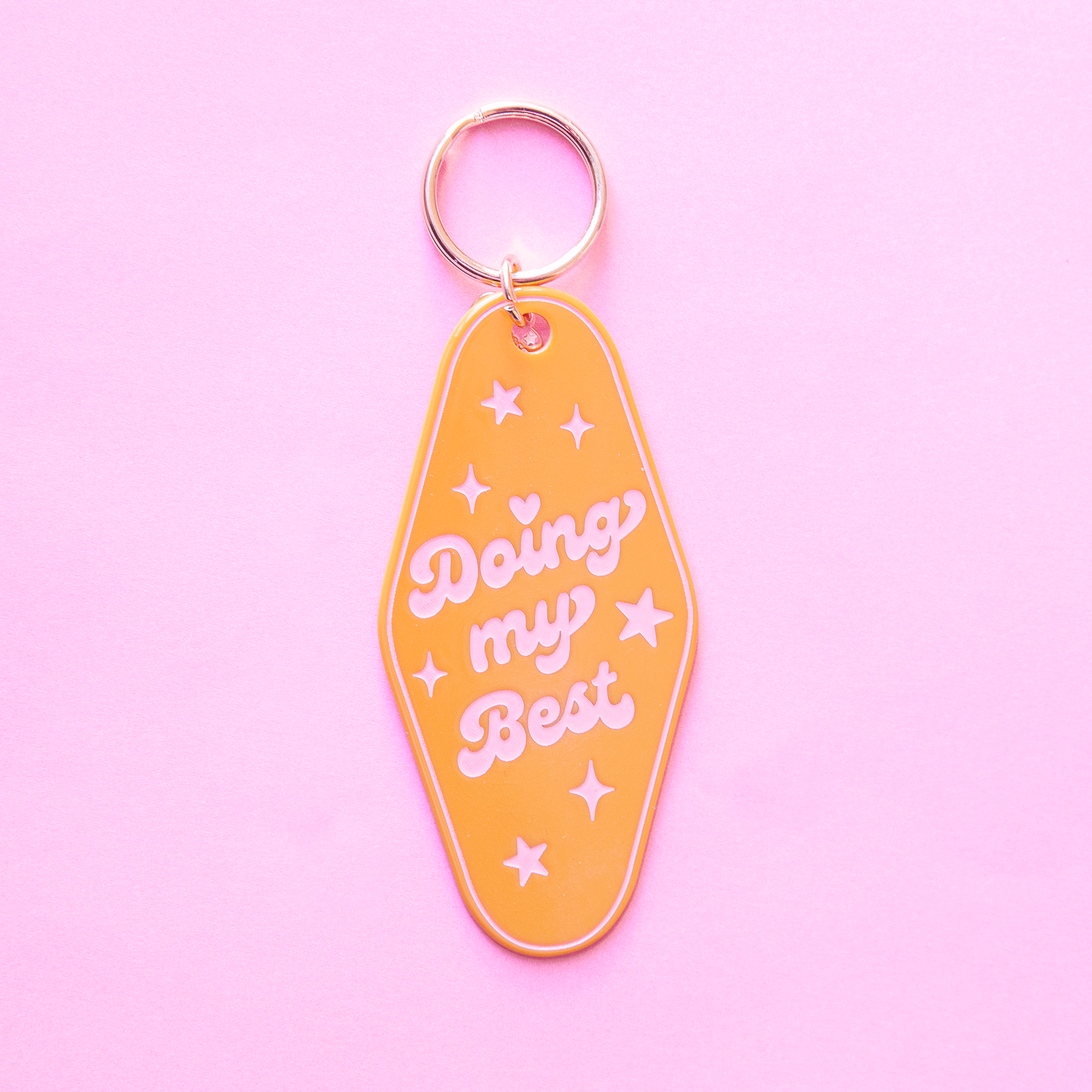 On a purple background is a mustard yellow diamond shaped keychain with a gold loop and pink text that reads, "Doing my Best" surrounded by star shapes and sparkles. 