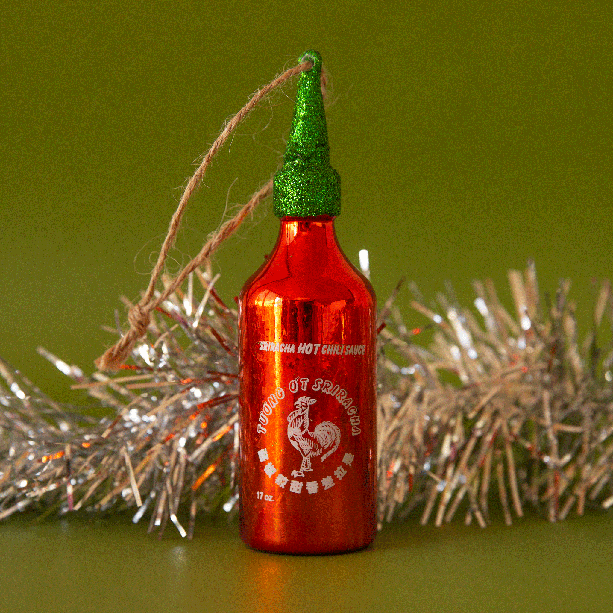 On a green background is a red and green glass sriracha bottle shaped ornament with the classic logo int he center and a twine loop for hanging. 