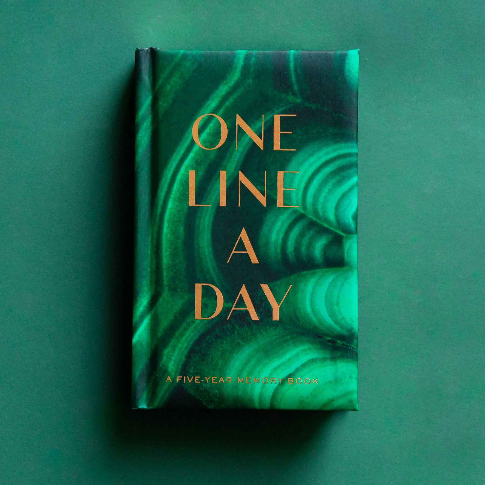 On a green background is a green malachite print journal with gold text that reads, &quot;One Line A Day A Five Year Memory Book&quot;.