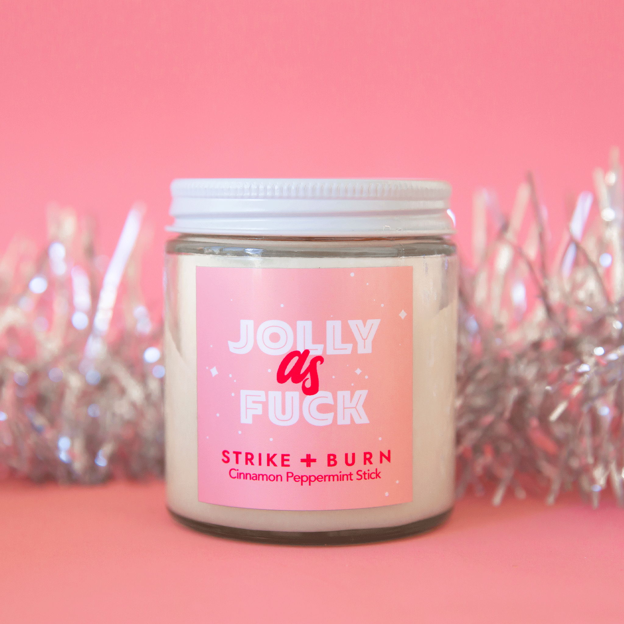A glass jar candle with a pink label that reads, "Jolly as Fuck" in white and hot pink font along with a white graphic of a Christmas ornament on the right side of the label.